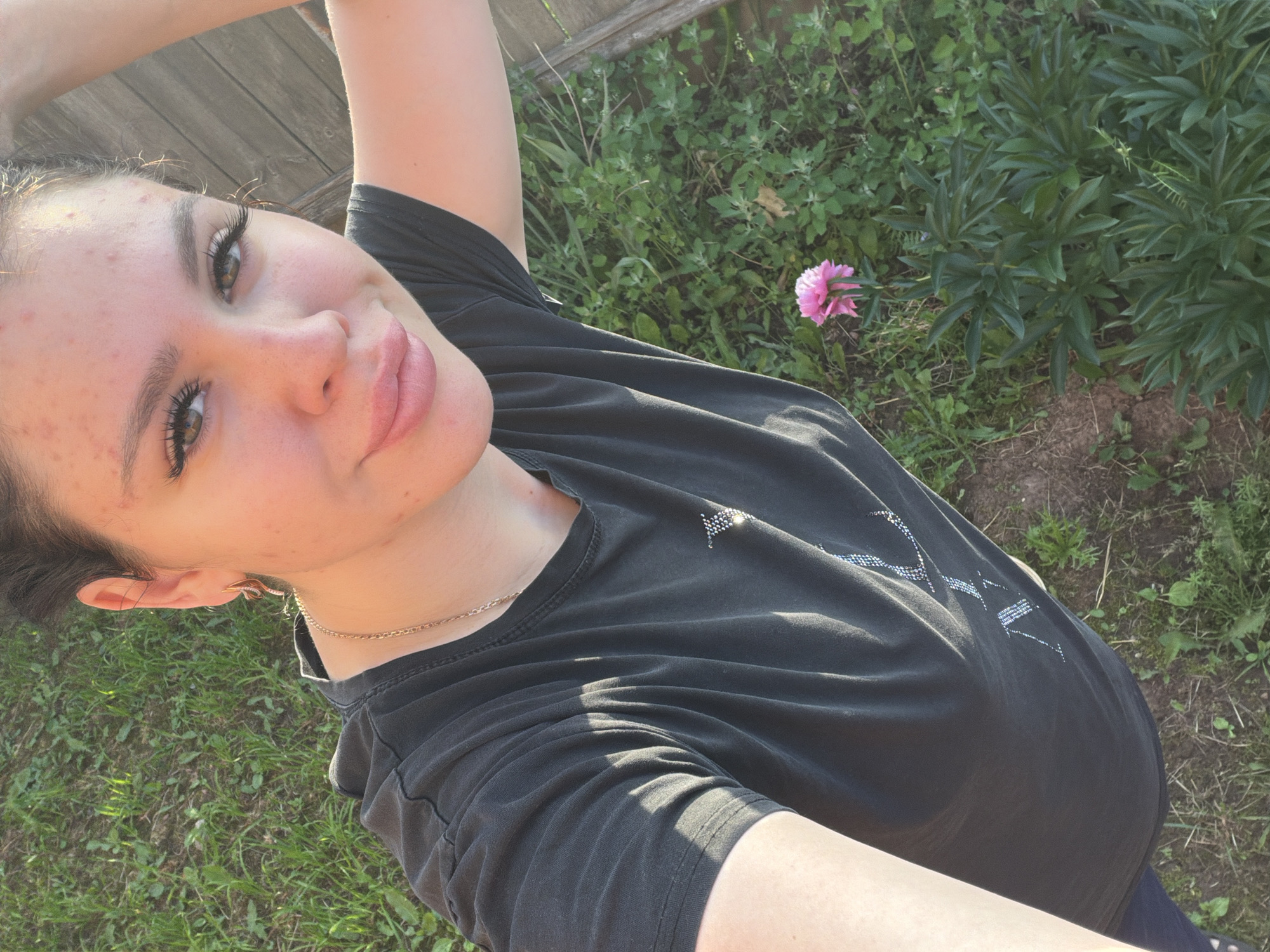 A young woman is taking a selfie outdoors, with a wooden fence and green grass in the background. She has fair skin, green eyes, and her dark hair is pulled back. She is wearing a black t-shirt with a silver chain necklace and dangling earrings. Her face has some acne. The lighting suggests it is late afternoon, with sunlight casting shadows on the grass and her t-shirt. In the lower right corner of the frame, a single pink flower is visible amidst green foliage. The setting appears to be a garden or yard, possibly in a rural or suburban area, consistent with a location in Bolshoy Bukor, Russia.