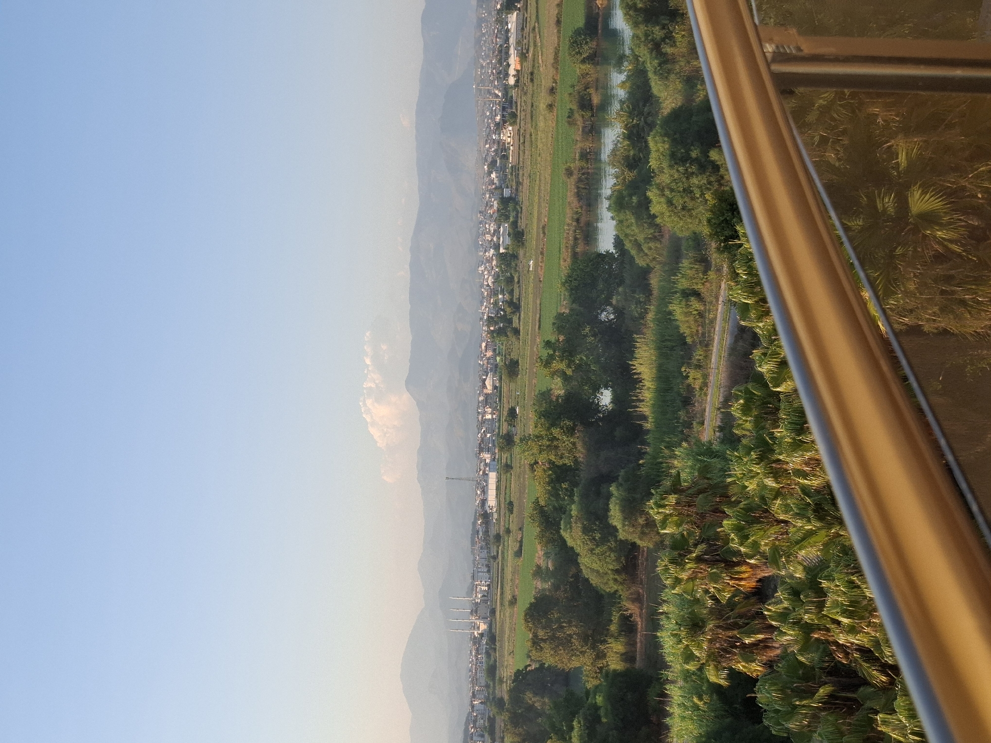 The image shows a panoramic view of Manavgat, Turkey, from an elevated position. In the foreground, a metallic railing and glass partition are visible, suggesting a balcony or viewing platform. Beyond this, dense green vegetation, including palm trees, dominates the middle ground. A body of water, possibly a river, is situated behind the trees, reflecting the sky. In the distance, a city sprawls across a plain, with buildings of various sizes and a prominent mosque with multiple minarets. A mountain range forms the backdrop, its peaks softened by the hazy atmosphere. The sky is a clear, pale blue, with a few wispy clouds. The overall impression is one of a sunlit landscape, likely captured during the day.