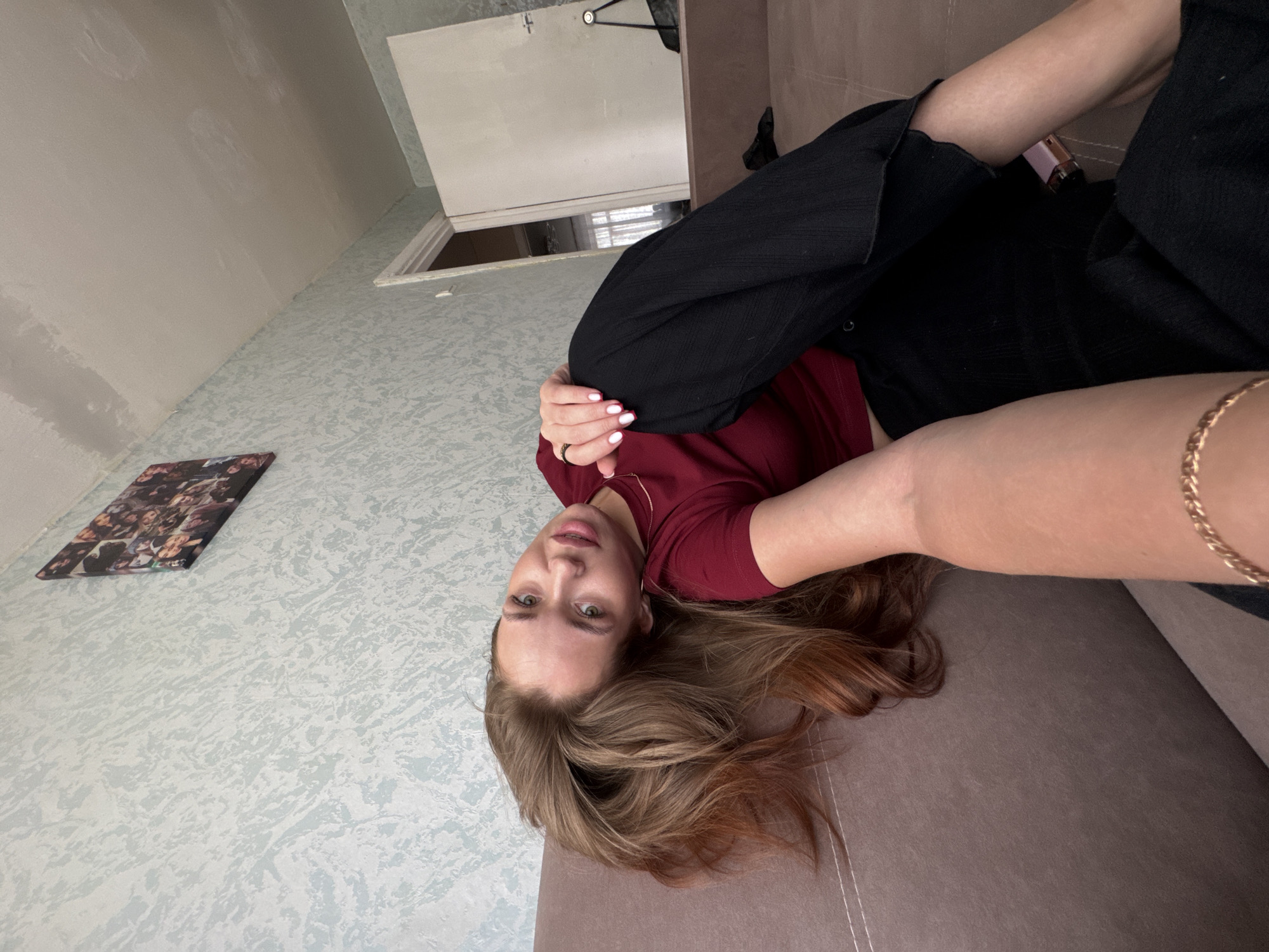 The image shows a young woman with long, wavy brown hair, wearing a burgundy t-shirt and black pants, reclining on a brown sofa. She is posing for a selfie, with her arm extended towards the camera. Her right hand, adorned with white nail polish and a ring, rests on her leg. The background features a light blue textured wallpaper and a door in the distance. A framed collage of photographs is visible on the wall. The setting appears to be an indoor residential space, likely an apartment in Chaikovsky, Russia, given the architectural style and decor.