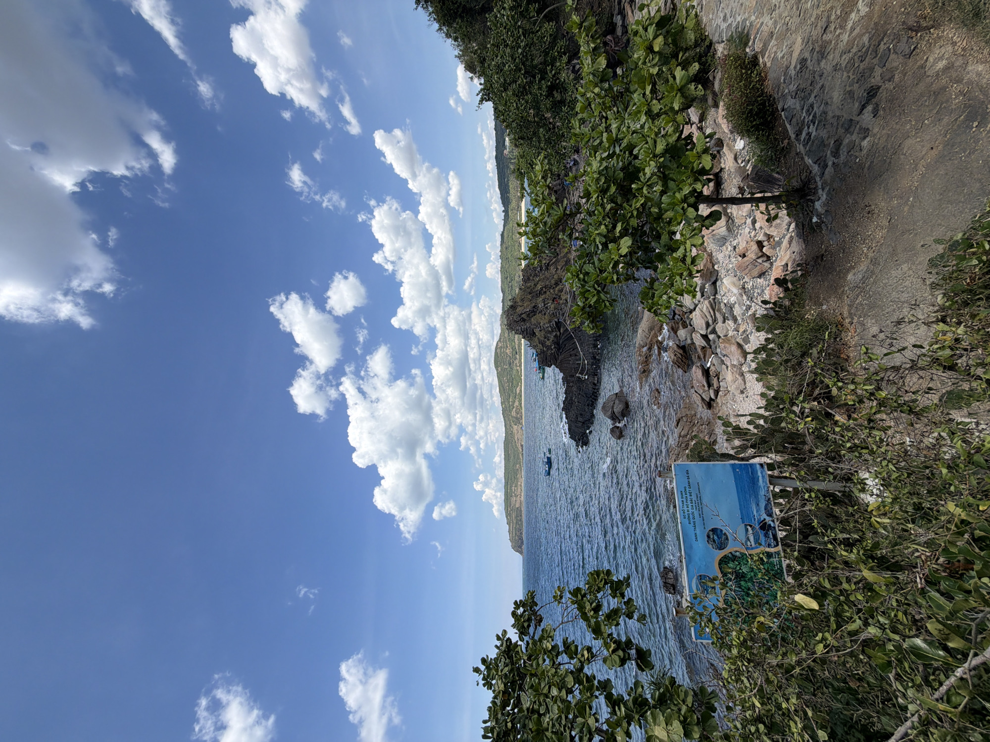 This is a landscape photograph taken from a rocky, vegetated area overlooking the sea. In the foreground, there are green trees and shrubs, with a path made of flagstones to the right. A sign with text and images is partially obscured by foliage in the lower left. The middle ground features the blue ocean with gentle waves, a rocky outcropping with dark, jagged rocks, and a few small boats. In the background, a coastal landscape with hills and more vegetation extends to the horizon under a bright blue sky with scattered white clouds. This scene is located in Phú Lương, Vietnam.