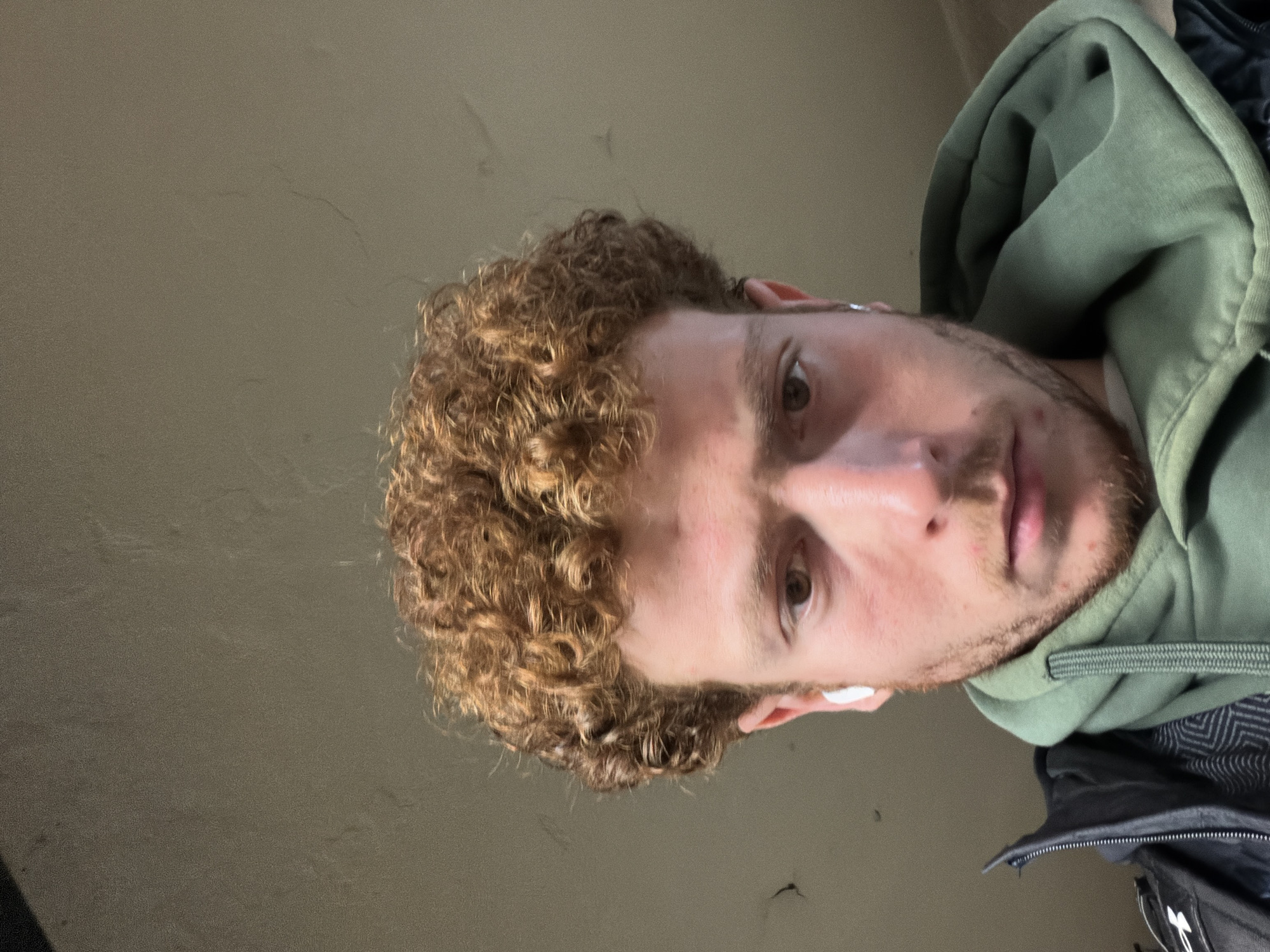 The image features a young man with curly, light brown hair and a light beard or stubble, looking directly at the camera. He has brown eyes and a neutral expression. He is wearing a green hooded sweatshirt with the hood up, and a dark jacket over it. A white earbud is visible in his left ear.
The photograph is taken from a low angle, looking up at the man, and is rotated 90 degrees clockwise, as if he is lying down or leaning back. The background is a plain, light brownish-grey wall or ceiling. There are no specific activities taking place, other than him posing for the photo. No text is visible in the image. Given the provided context, the setting is in New York, United States of America. The time of day and weather are not discernible due to the plain indoor-like background and lighting.