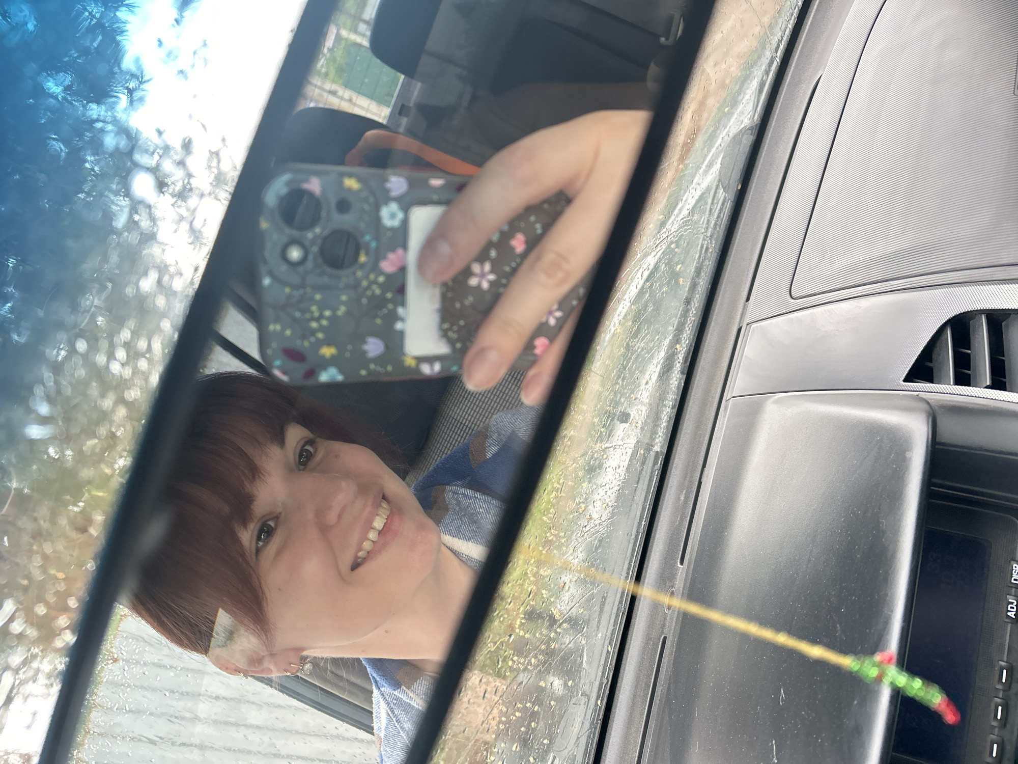 A woman is taking a selfie in the rearview mirror of a car. The woman has brown hair with bangs and is wearing a blue and white plaid shirt. She is smiling and holding up her phone, which has a floral case. The rearview mirror is reflecting the woman's face and the interior of the car. The dashboard of the car is visible, and there is a decorative string hanging from the rearview mirror. It is raining, and the windshield is covered in water droplets. The location is possibly Фоки, Russia.