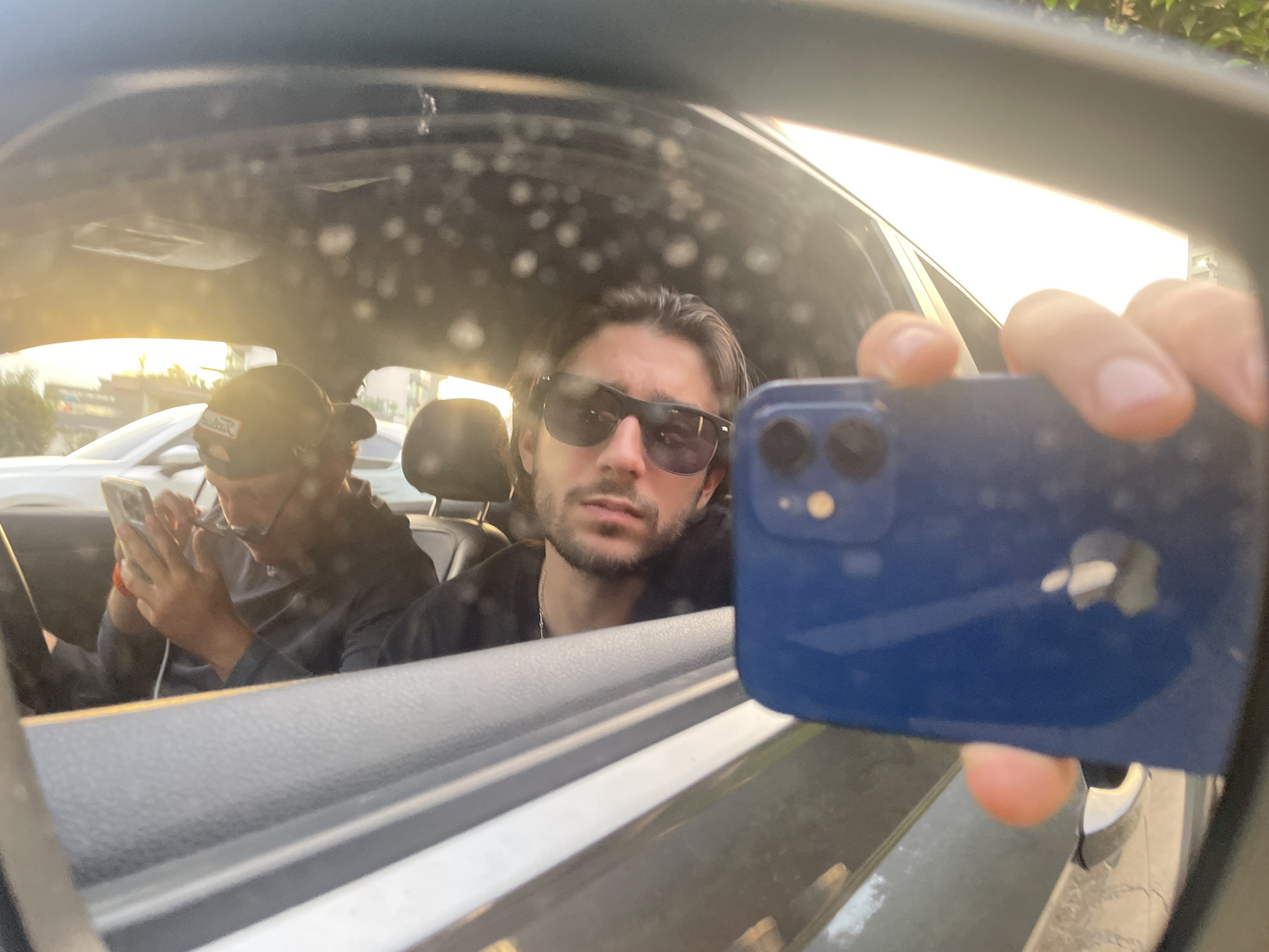 Captured through a car's side mirror, a man with sunglasses in a black top is seen holding a blue smartphone, presumably taking the picture. Inside the car, another individual wearing a baseball cap is focused on their own smartphone. The scene appears to be in an urban setting, illuminated by sunlight. While the city is stated as Huixquilucan de Degollado, Mexico, there are no details confirming that.