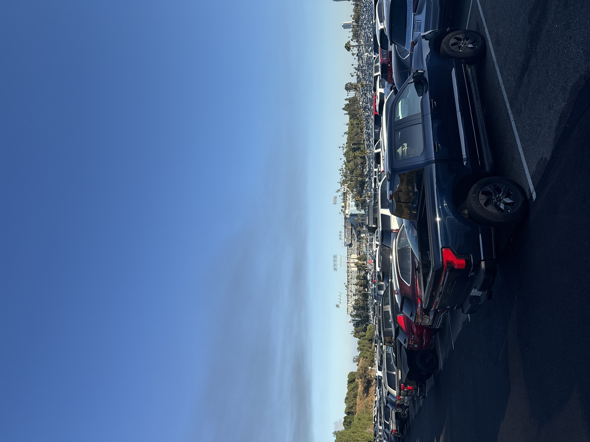 This is a wide-angle, eye-level shot of a vast parking lot filled with cars, extending towards a stadium in the distance, under a clear blue sky. The foreground is dominated by a dark blue pickup truck, a Ford F-150 Lightning, parked in a marked space. The truck's taillights are illuminated. Behind it, other vehicles are visible, including SUVs and sedans in various colors, such as red, white, and gray.
In the middle ground, rows upon rows of cars fill the parking lot, creating a sea of vehicles. Beyond the parking lot, a sprawling cityscape is visible, with a large stadium complex featuring multiple towers and light poles. Palm trees are scattered throughout the landscape, and a distant skyscraper can be seen on the right side of the image. The background features a hazy horizon, suggesting the presence of smog or smoke. The overall impression is that of a large event or venue, with a significant number of attendees having arrived by car.
Based on the presence of the stadium and the overall landscape, this image was likely taken at Dodger Stadium in Los Angeles, California. The stadium is a prominent landmark in the Elysian Park area of Los Angeles, known for its large parking lots that accommodate thousands of vehicles for games and events. The surrounding hills and vegetation are also characteristic of the Los Angeles basin.