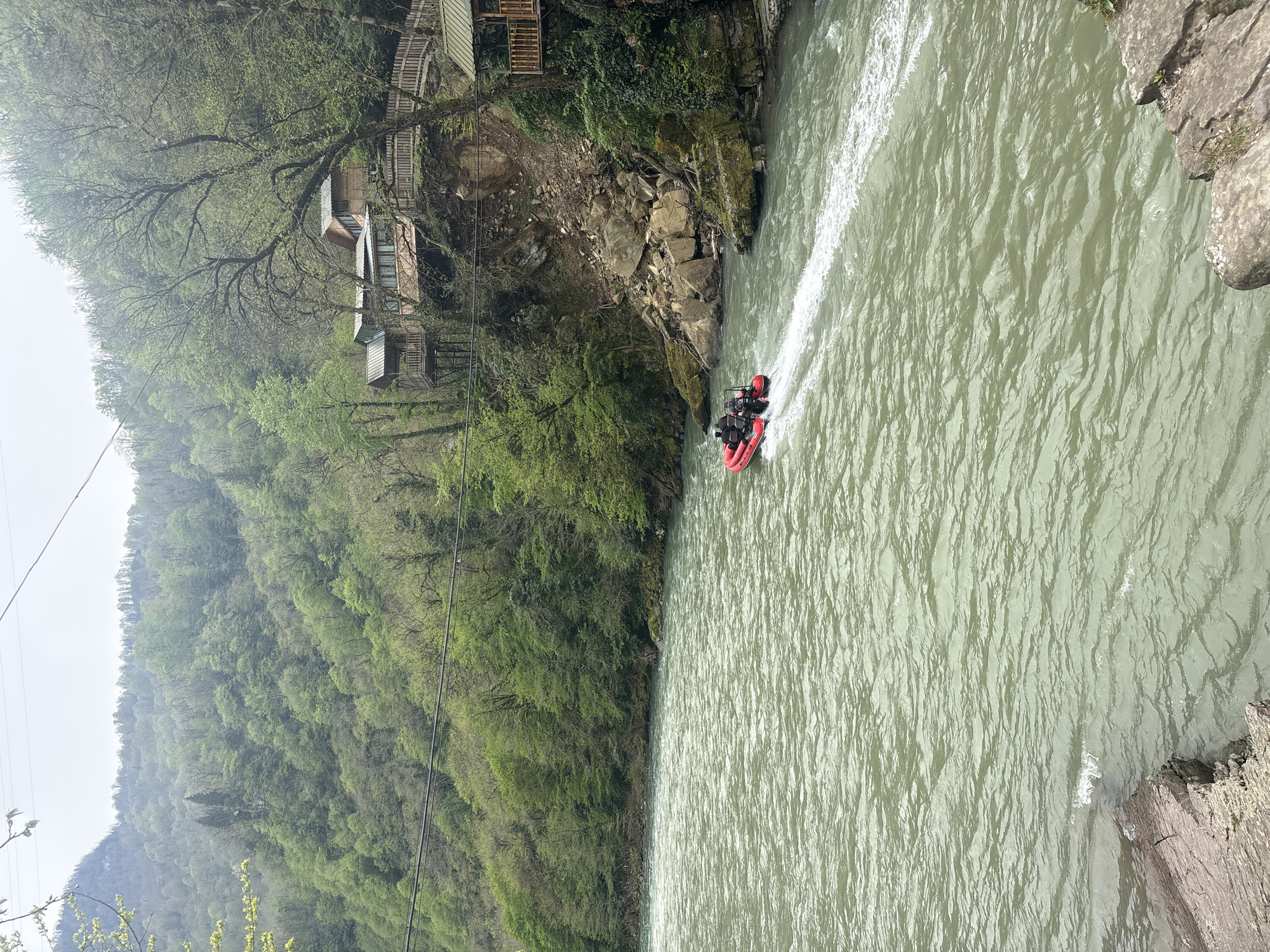 The image shows a red inflatable boat in a river at Kveda Makhuntseti, Georgia. The boat is traveling rapidly through the water, creating a white wake behind it. On the left bank of the river, there's a dense forest covering a steep hillside. Higher up the hill are a few wooden buildings constructed on stilts, nestled amongst the trees. The river's water is a murky green color. The right bank is rocky. The sky is overcast. Overhead power lines are visible.