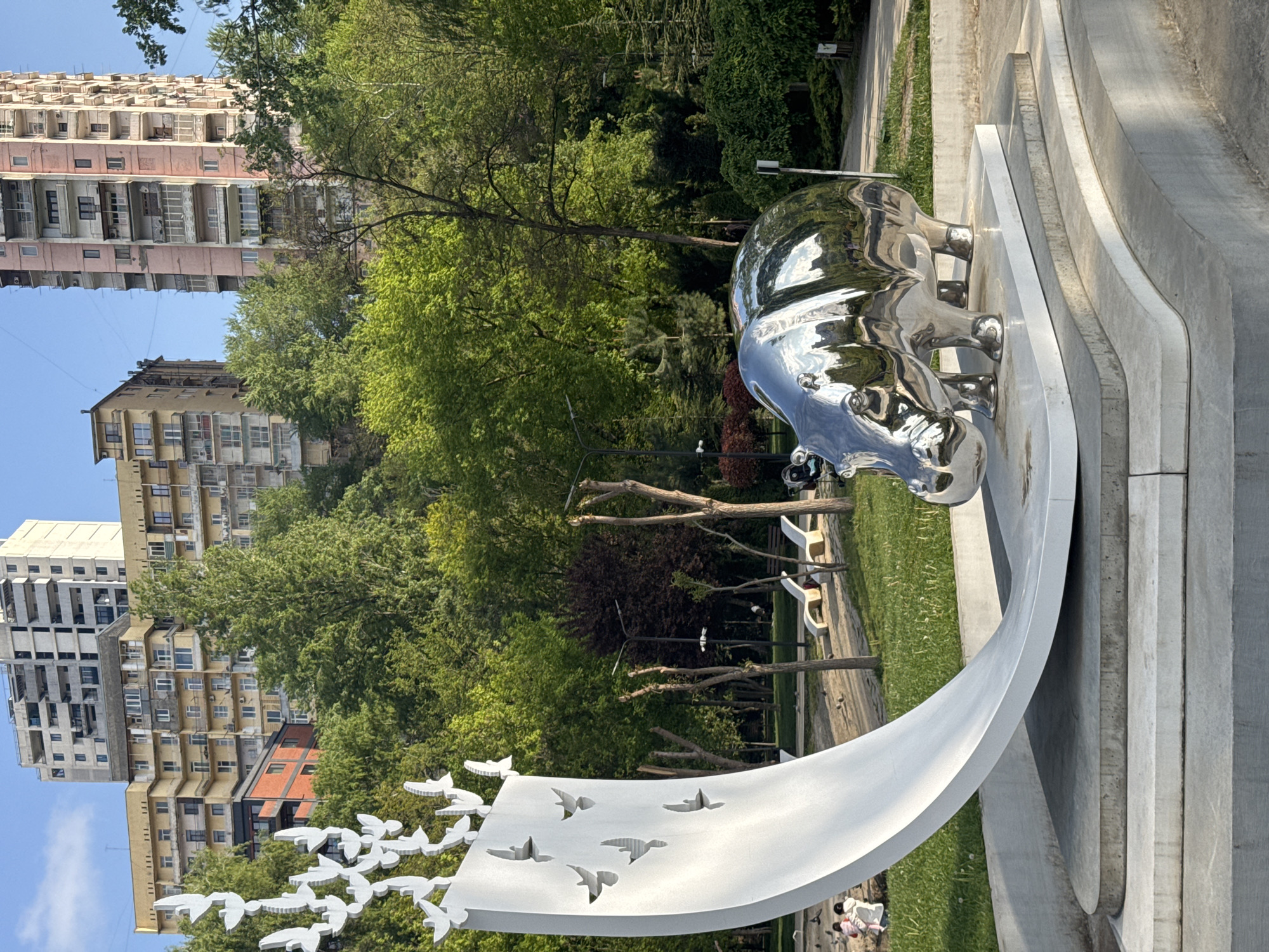 A vertically oriented outdoor photograph captures a park scene featuring modern sculptures against an urban backdrop. In the foreground, a large, highly reflective, silver-colored sculpture of a rhinoceros stands on a white, curving base. The rhino's metallic surface gleams, reflecting the surrounding trees and sky. To the left of the rhino, a white, abstract structure curves upwards, featuring numerous white bird or dove figures emerging from or cut into its surface, giving the impression of flight.
The sculptures are situated on a well-maintained grassy area. Behind them, a dense cluster of green trees creates a natural buffer. Through the foliage and above the treeline, several multi-story residential buildings are visible in the background, characterized by numerous balconies and a somewhat dated architectural style, suggesting apartment blocks. Some buildings are light pink or beige, while others are gray. A clear, bright blue sky is visible above the buildings.
Subtle hints of activity include a paved pathway winding through the park and a few benches. A small group of people, including children, are faintly visible near the base of the white dove sculpture, appearing to relax or interact with the artwork. The scene is bathed in bright daylight, indicating clear, sunny weather, possibly mid-morning or afternoon given the light and shadows. No discernible text is visible in the image.