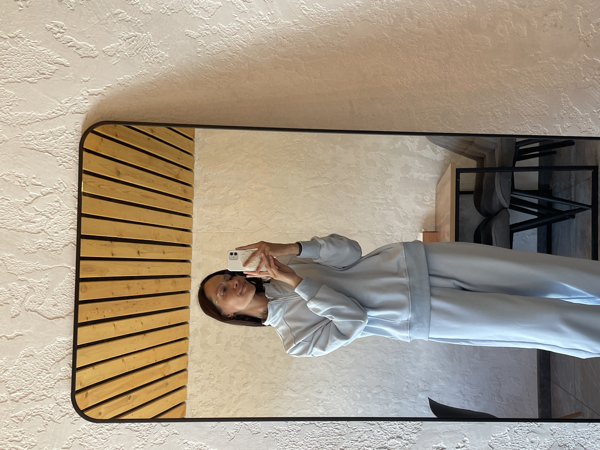 In Oktyabrsky, Russia, a young adult female takes a mirror selfie in an indoor establishment, likely a cafe or lounge. The woman, with long dark brown hair and light-rimmed glasses, wears a light blue full-zip tracksuit consisting of a long-sleeved sweatshirt with visible seams and loose-fitting trousers. She holds a white smartphone with a textured light-colored case in both hands, positioned to capture her reflection in a large, black-framed mirror with rounded upper corners. A subtle smile is present, and a small tattoo is visible on her neck.
The mirror reflects a light-colored wooden slatted ceiling arranged diagonally and a prominent, off-white, heavily textured wall. Also visible in the reflection are a light wood-topped table with a black metal frame and two dark gray upholstered chairs with black legs. The floor inside the reflection is dark. Outside the mirror, the same off-white, textured wall extends, and a segment of a black chair is visible to the far left. The setting features a modern interior aesthetic.