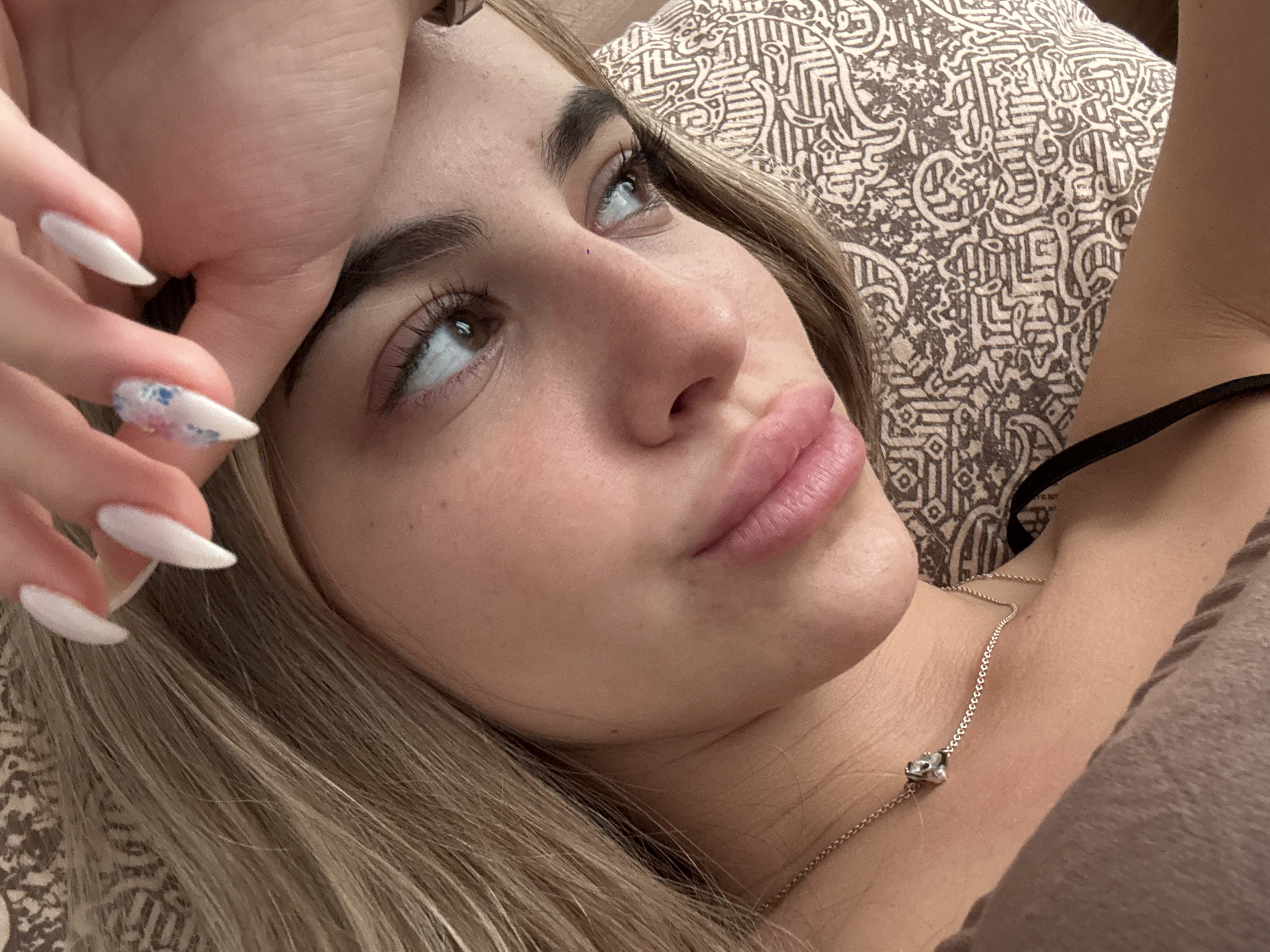 A close-up portrait captures a young woman lying down in an indoor setting, likely a bedroom. She looks upwards and slightly to her right, her expression appearing contemplative or pensive. Her right hand rests gently on her forehead, revealing long, pointed nails, predominantly white, with one nail featuring a subtle blue and pink watercolor-like design.
The woman has light-colored (blonde or light brown) hair, parted and falling around her shoulders. Her features include full, naturally pink lips, brown eyes with long lashes, and well-defined eyebrows. Around her neck, she wears a delicate silver chain necklace with a small, reflective pendant, possibly a heart or an abstract charm, resting on her décolletage. A thin black strap from her top is visible near her shoulder.
The background shows a patterned pillow or cushion on the right side of the frame, featuring an intricate brown and beige geometric design. On the left, a plain, light brown fabric, possibly her clothing or bedding, completes the scene. The lighting is soft and even, creating a gentle and intimate atmosphere. There are no specific visual cues within the image to determine the time of day or external weather conditions. No visible text is present. The provided context indicates the location is Sofia, Bulgaria.