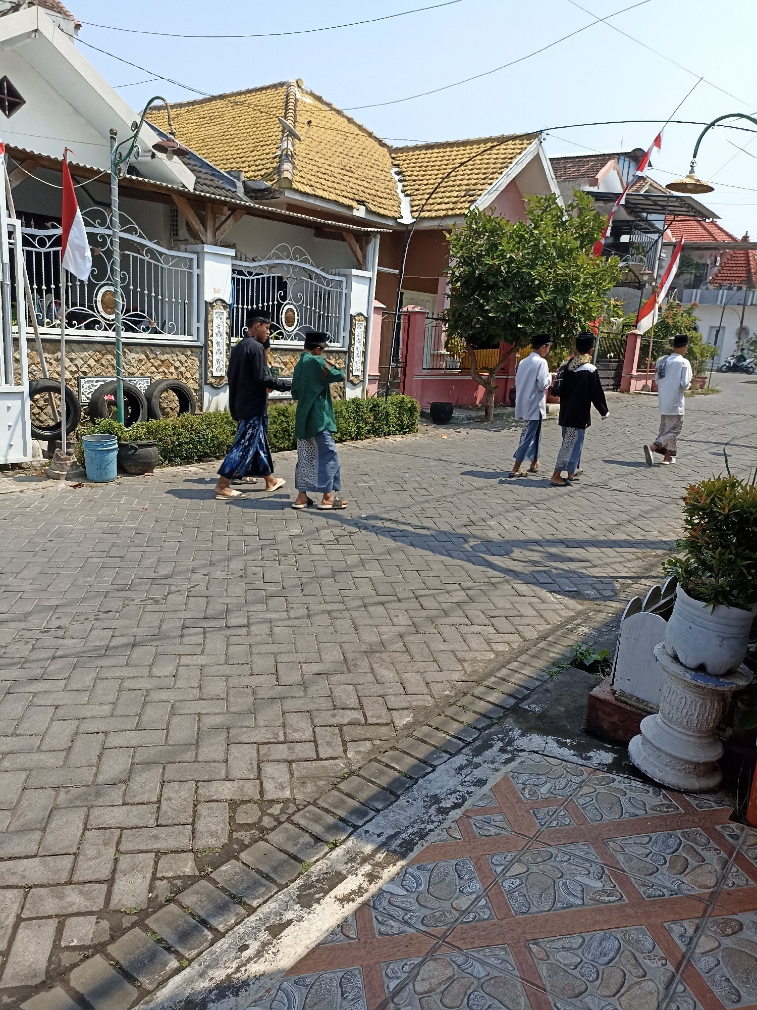 In Sumberejo, Indonesia, on a clear day with a bright blue sky, five young males are walking on a brick-paved road. They are all wearing traditional Indonesian attire consisting of sarongs and shirts, with some also wearing caps. Two of the individuals in the foreground are walking side-by-side, while the other three are further down the road, also walking in the same direction. The scene is set against a backdrop of residential houses with orange tile roofs, some adorned with Indonesian flags. To the left of the frame, a white fence with intricate metalwork encloses a property, and several used tires are stacked near the entrance. A blue bucket is visible next to the tires. The road is lined with green shrubs and trees. In the background, a motorcycle is parked. The foreground of the image shows decorative tiled flooring bordering the brick pavement.