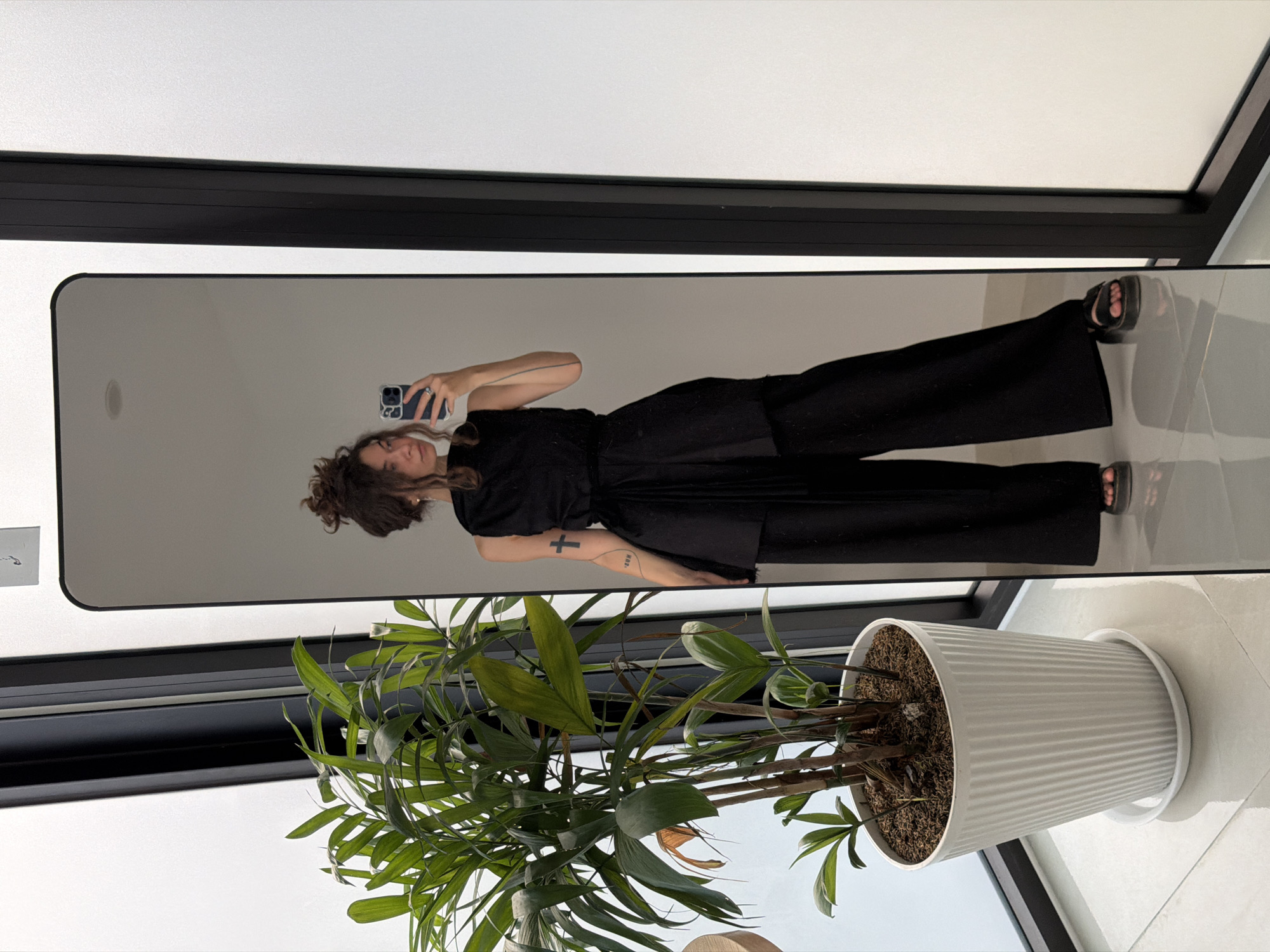 A full-length mirror selfie captures a person, likely female, standing indoors in a modern setting in Đà Nẵng, Vietnam. The person has dark, curly hair styled in a bun and is wearing a black short-sleeved top, wide-leg black trousers, and black open-toed sandals. She holds a dark-colored smartphone to take the photograph, displaying a slight smile. Notable details on her left arm include a small black cross tattooed on the inner forearm and a word, possibly "SUN", tattooed further up. A ring is visible on her right hand.
The mirror, framed in black, reflects a brightly lit interior. To the right of the mirror, a large white ribbed planter holds a lush green leafy plant, resembling a palm. The floor is light-colored, possibly white or light grey tiles. Beyond the immediate foreground, a large window or glass door with a black frame allows ample natural light into the room, indicating it is daytime. A black horizontal beam structure is visible across the top of the image. The scene suggests a relaxed and casual moment.