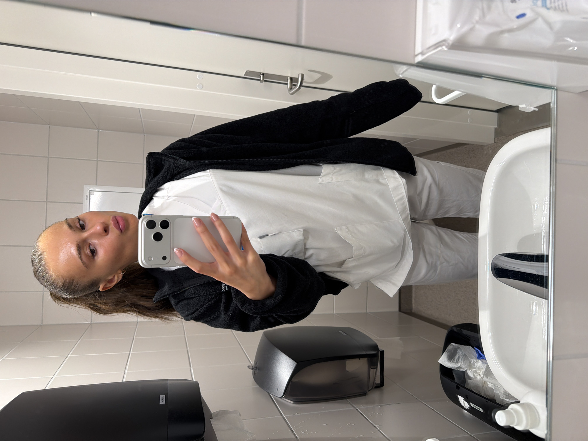 The image shows a young woman, likely a healthcare professional, taking a mirror selfie in a public restroom. She is wearing white scrubs and a black zip-up jacket. Her hair is pulled back in a high ponytail, and she is making a kissing face. The restroom appears to be in a hospital or clinic setting, with tiled walls, a paper towel dispenser, a trash can, and a sink visible in the reflection. The lighting is bright, and the overall impression is one of a casual moment captured during a workday. The context of Harstad, Norway, is not directly evident from the visual information within the image itself.