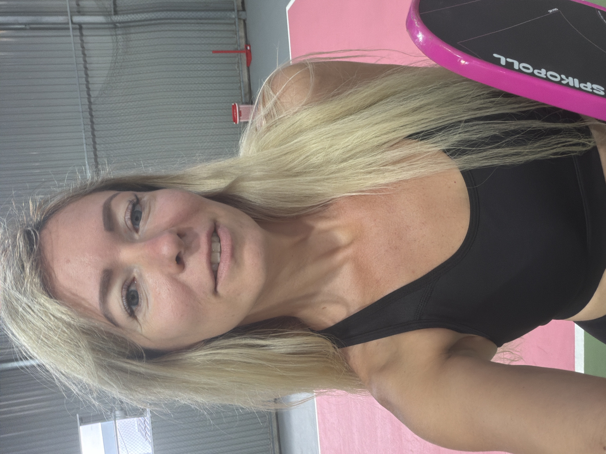A selfie-style, medium-shot photograph shows a blonde woman with fair skin and light blue eyes. She is wearing a black sports bra and appears to be indoors, possibly in a sports facility with a gray corrugated metal wall in the background. A pink and black paddle is held in her right hand, with "SPIKOPOLL" visible on it. The floor appears to be a court with pink and green markings. The woman is looking directly at the camera with a slight smile.