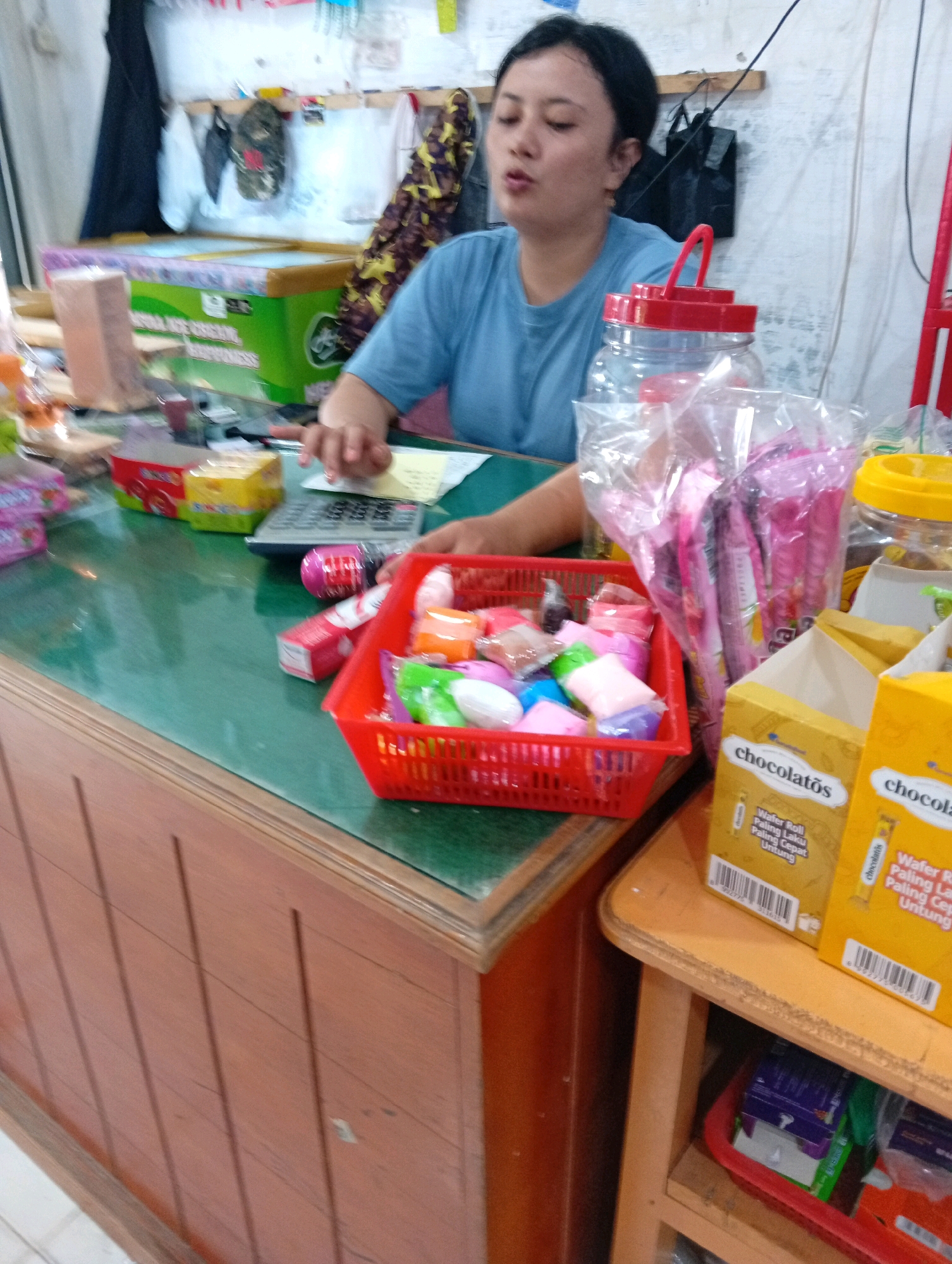 The image depicts a retail setting, possibly a small convenience store or a warung, in the City of Gunungsitoli, Indonesia. In the foreground, a wooden counter with vertical planks is visible. On the counter, a green surface is covered with various items.
To the left, there is a green and white chest freezer with text that is partially obscured but appears to relate to ice cream. Next to it are some cardboard boxes and a brown object. Further to the right on the counter, a calculator is placed next to a person's hands. The person, a woman with dark hair pulled back and wearing a blue t-shirt, is seated and appears to be interacting with the calculator and possibly engaging in a transaction or inventory. Her facial expression suggests concentration.
In front of the woman and to the right, a red plastic basket holds an assortment of colorful, individually wrapped items, likely candies or small toys. Behind the basket, several plastic bags filled with pink packaged goods are stacked, possibly snacks or confectioneries. To the far right, there are two yellow boxes with the brand name "Chocolatos" printed on them, indicating they contain wafer rolls. A yellow plastic container with a yellow lid is also visible.
The background shows a wall with items hanging on it, including bags, clothing, and what appears to be a camouflage-patterned hat. The overall environment suggests a modest, functional retail space. The presence of the chest freezer, the variety of packaged goods, and the counter setup strongly suggest a local shop selling everyday consumer items.