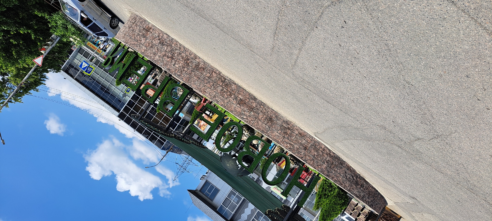 The image shows a street scene in Yessentuki, Russia. The focal point is a large, green, artificial grass sign spelling out "HELLO!" in large letters. Behind the sign is a modern building with a glass facade. A silver car is parked on the right side of the image, and a pedestrian crossing sign is visible on the left. The sky is blue with scattered white clouds, and trees are visible in the background. The photo is taken from a low angle, with the street in the foreground.