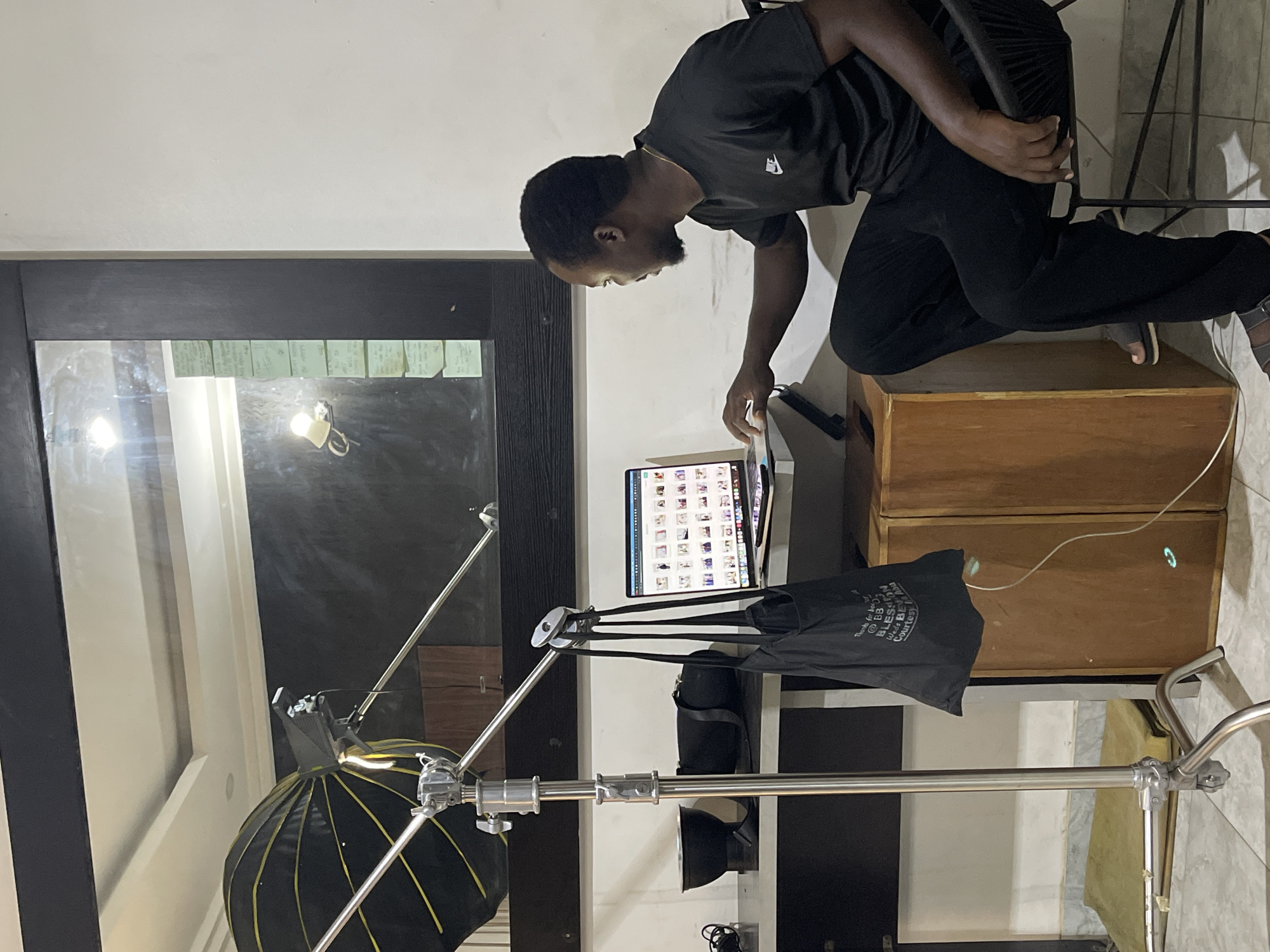The image depicts a man, likely in Abuja, Nigeria, engaged in photo review or editing within an indoor, studio-like setting. The man, with dark hair and a beard, wears a black t-shirt with a white logo (resembling a Nike swoosh) and black trousers. He is bent forward, focused on a laptop or large tablet screen that displays a grid of thumbnail images. He is seated on a black chair, which is elevated on two stacked wooden boxes acting as a workstation.
To his left, professional photography equipment is visible, including a partially collapsed black softbox with yellow ribs, mounted on a sturdy silver light stand. Another silver light stand is in the foreground. A black tote bag hangs from one of the light stands, featuring white text that reads: "Thanks for trusting @BBL_NIGERIA where Beauty is a duty".
The background includes a white wall and a dark-framed surface, possibly a mirror or window, which reflects bright artificial light sources and several green sticky notes. The floor is tiled in a light grey. The scene is artificially lit, suggesting an indoor workspace. The man appears attentive and concentrated on his task.