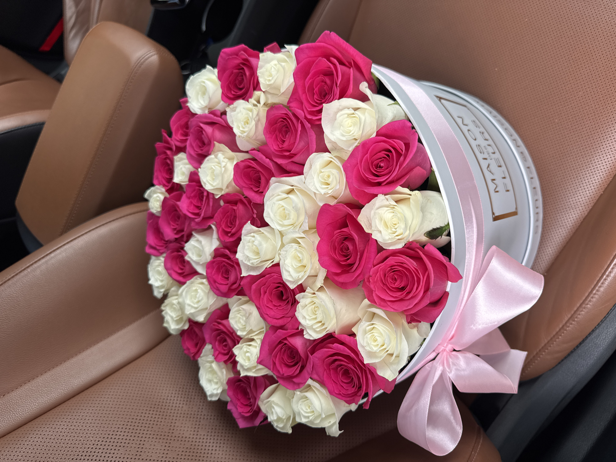 A bouquet of white and fuchsia roses is arranged in a white circular box tied with a pink satin ribbon. The box sits on the brown leather interior of a car. The roses are full and varied in bloom, with some tightly budded and others fully open. The contrast between the deep fuchsia and pure white petals is striking. The box has text printed on its side, partially visible, which reads "MAISON DES FLEURS". The background shows the textured brown leather seats of the car, suggesting a luxurious and private setting. The lighting is soft, indicating it could be daytime, though the exact time is not discernible. The overall impression is one of elegance and a thoughtful gesture.
