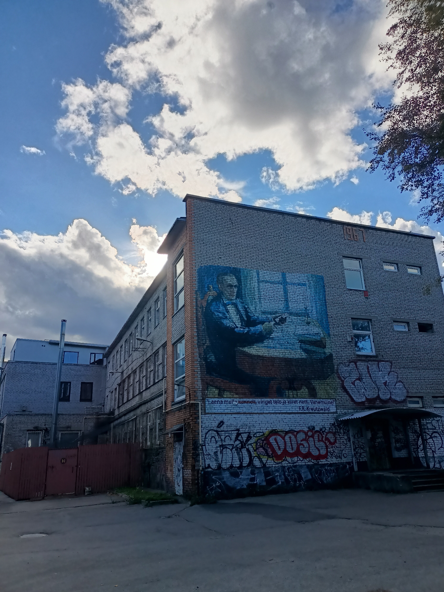 This is a medium-shot, eye-level, outdoor, daylight photograph taken in Tallinn, Estonia. The image features a brick building with a large mural on its side depicting a man in a blue suit sitting at a table, seemingly engaged in writing or composing. The mural is set against a blue background and extends across a significant portion of the building's facade. Below the mural, there is graffiti on the brick wall, including stylized lettering. The building has multiple windows, some of which reflect the sky and clouds. To the left of the main building, another, lighter-colored brick building is visible, with a red wooden fence in front of it. The sky above is partly cloudy with bright sunlight breaking through the clouds. In the upper right corner, the dark branches of a tree are silhouetted against the sky. The foreground consists of a paved area, likely a courtyard or parking lot. The overall impression is of an urban environment with elements of street art and architecture.