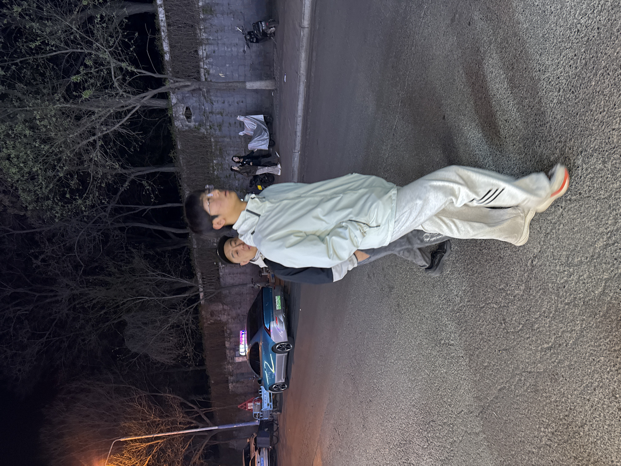 This media file captures a nighttime street scene in Zhongtun, China, featuring two young men in the foreground. The taller man, positioned slightly ahead, wears a light-colored jacket, light grey sweatpants with black stripes, and white-soled sneakers with orange accents. He has short dark hair and glasses. Partially visible behind him is another man with short dark hair, wearing a dark jacket or hoodie and dark pants. Both men are standing on what appears to be an asphalt or concrete road surface.
In the mid-ground, other individuals are seen near a silver tarp-covered electric scooter parked against a low stone wall or fence. Another dark-colored scooter or motorcycle is parked further along the wall. A light blue car, possibly an electric vehicle, is parked closer to the foreground. Its license plate is visible, displaying "京 A 51840 绿" (Jing A 51840 Green), indicating a Beijing license plate for a new energy vehicle. Partially obscured Chinese text, including the character "急" (jí - urgent/emergency), is visible on a sign behind the car.
The background consists of dark, bare-branched trees, with some showing sparse new leaves, silhouetted against the dark night sky. A street light with an orange glow illuminates parts of the scene from the left. The overall lighting suggests it is late evening or night. The mood is neutral, with no overt activity or strong emotions displayed by the subjects.