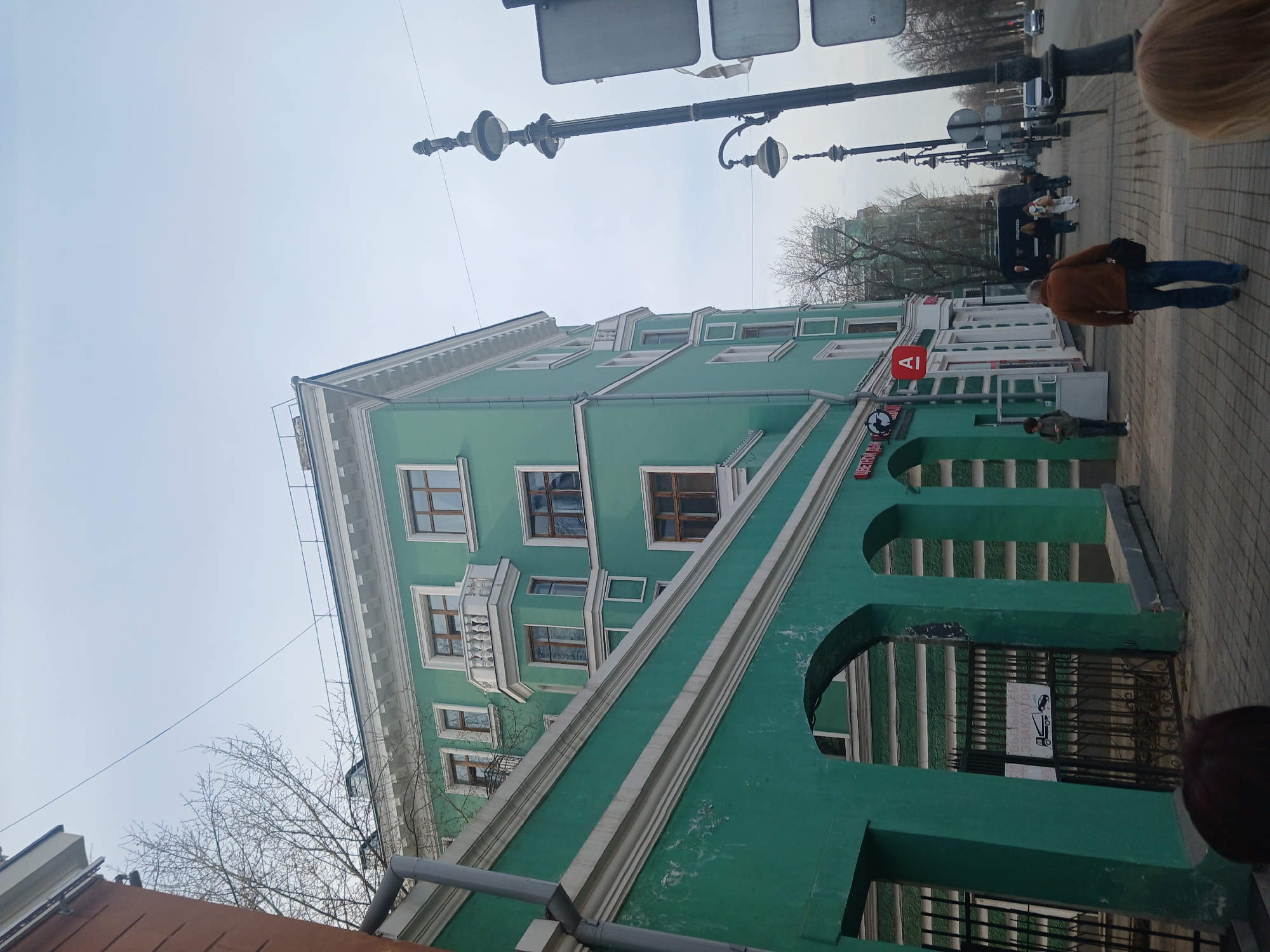The image captures a street scene in Perm, Russia, featuring a prominently green, multi-story building with white architectural details, including window frames, balconies, and an arched entryway. The building appears to be a historical structure. A red and white sign is visible on the facade. Pedestrians are seen on the sidewalk, with one person walking away from the camera and another standing near the building's entrance. Street lamps line the street, and a few vehicles are visible further down. The sky is overcast. A section of what appears to be a building's roof or canopy is in the lower left corner of the shot. The shot is from a slightly low angle.