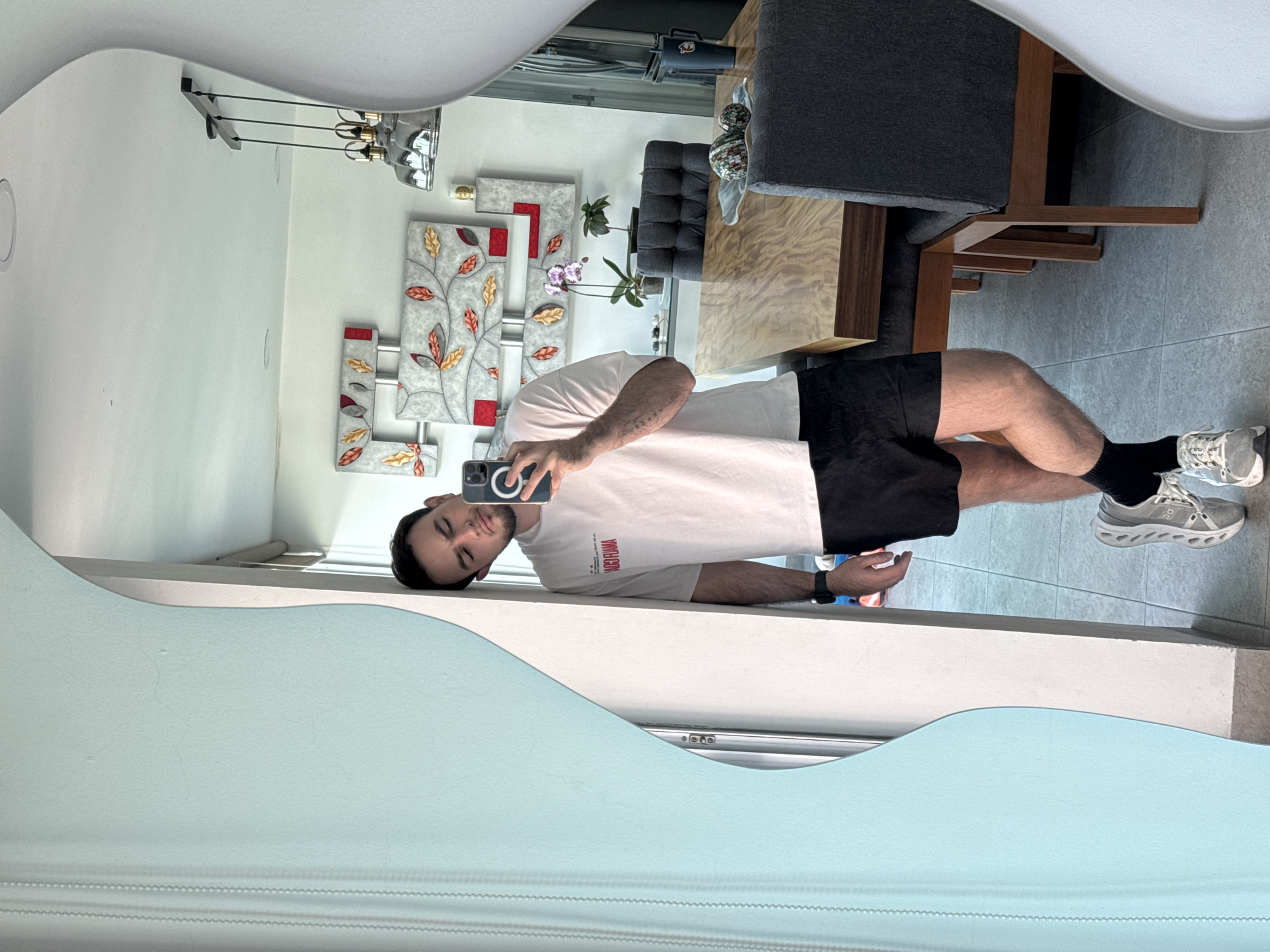 A person is taking a photo of themselves in a mirror inside a room. The person is wearing a white T-shirt and black shorts, along with black socks and gray running shoes. The room contains decorative wall art of leaves, a gray chair, and a wooden table. Blinds are visible in the mirror's reflection, and the person's reflection also shows part of a window or wall. It is not possible to independently verify the location is San Bernardino Tlaxcalancingo, Mexico, from the provided image.