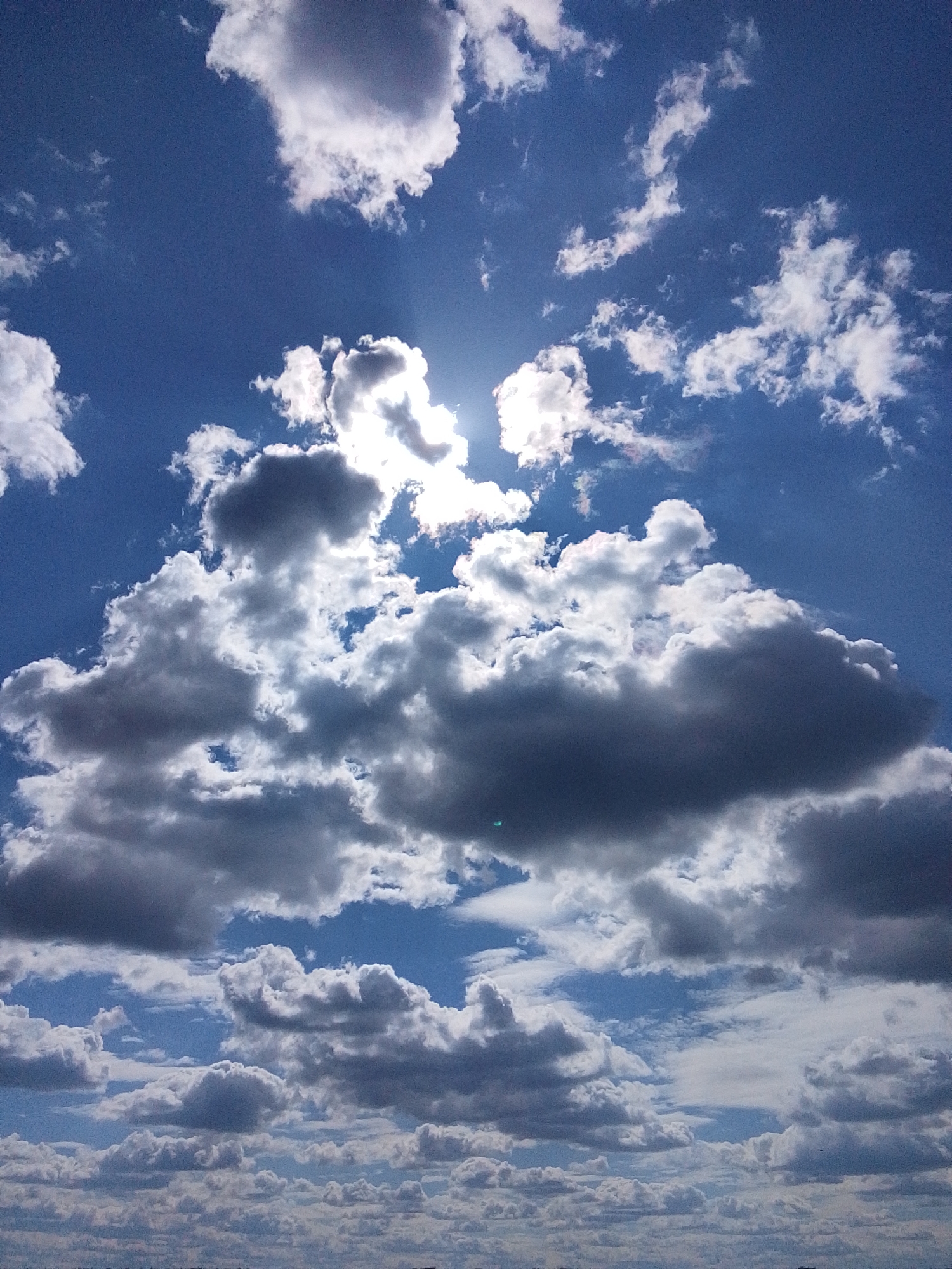 The image is a view of the sky with various types of clouds. The sky is blue, and the clouds range from small, puffy cumulus clouds to larger, darker cumulonimbus clouds. The sun is visible, partially obscured by clouds. The location of the image is identified as Khlevnoye, Russia. The image contains no people, objects, actions, or interactions. The environment is dominated by the sky and clouds.