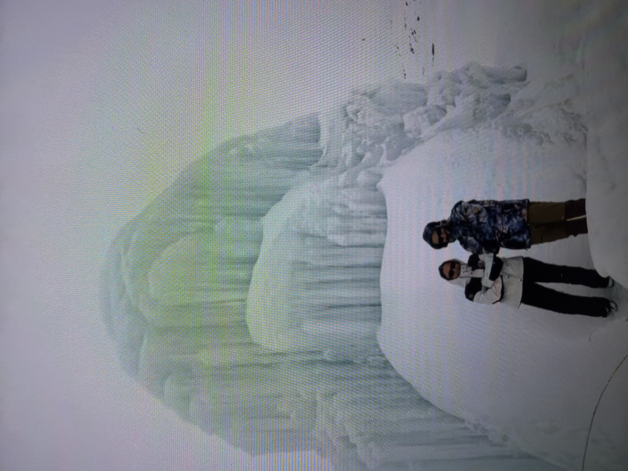 Two individuals stand on a snow-covered surface near a large ice formation. One person wears a patterned jacket and brown pants, while the other wears a light-colored jacket with black pants. The scene appears to be outdoors in a cold climate. Location is Unknown, Unknown.