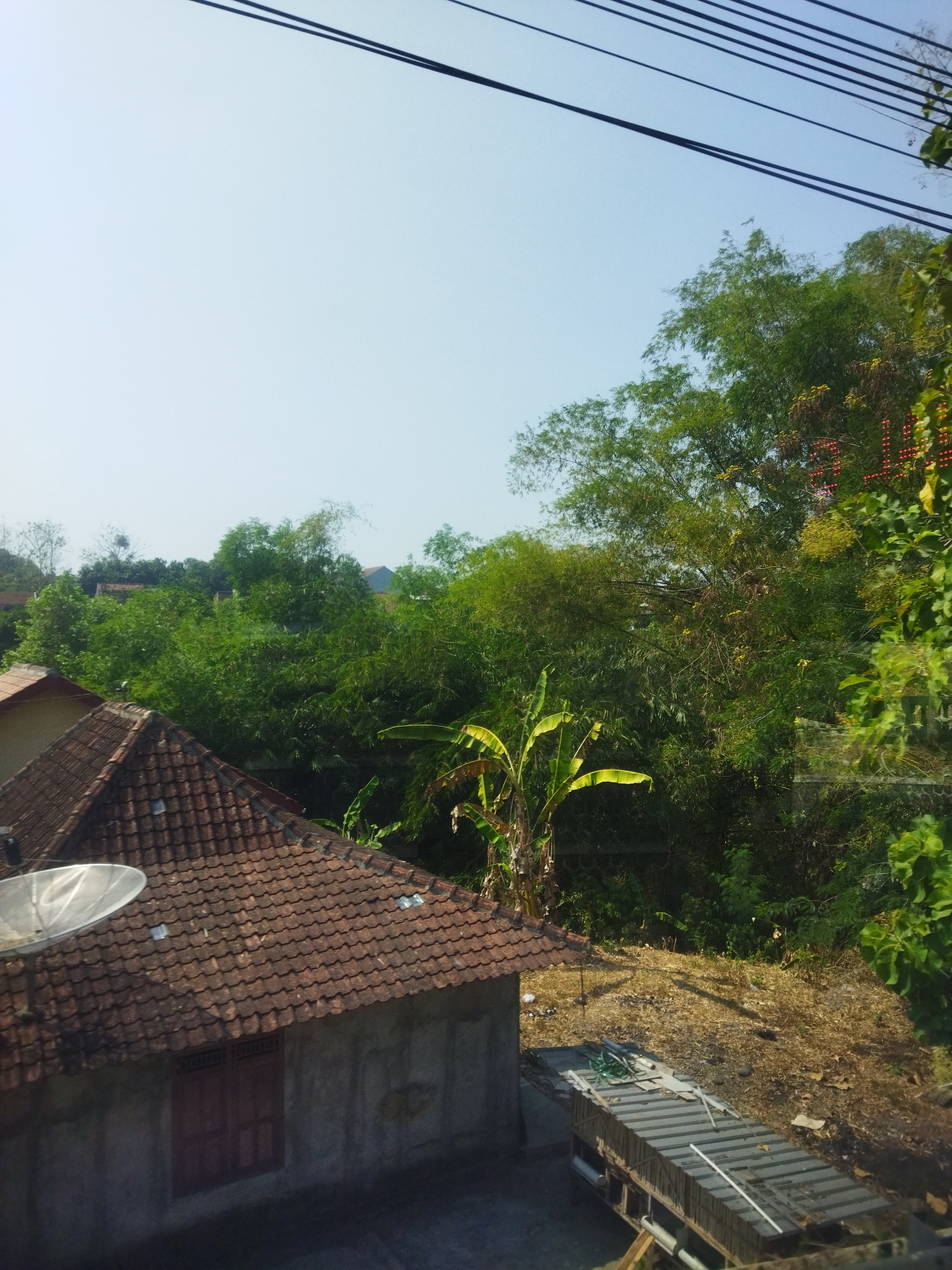 The image displays a view from a window, looking out over a residential area. In the foreground, there is a portion of a house with a tiled roof and a satellite dish. Adjacent to it, a small structure with a sloped, corrugated metal roof sits on a wooden frame. Beyond these structures, a dense area of green foliage, including bamboo and banana trees, dominates the midground. The background shows a clear blue sky with several overhead power lines. The scene is likely in a tropical or subtropical region. The location is identified as Singodutan, Indonesia.