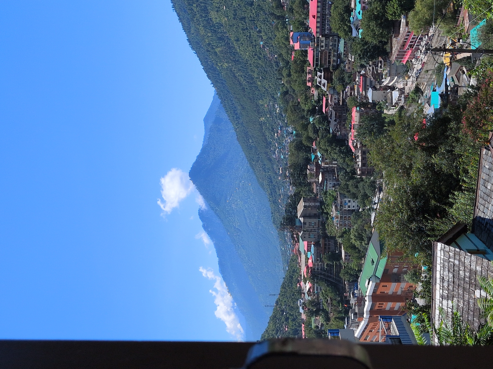 A medium-shot, elevated view captures the city of Kullu, India, nestled within a valley and framed by lush, forested mountains under a bright blue sky dotted with sparse white clouds. In the foreground, a dark, vertical element on the left, possibly a window frame or structural beam, partially obscures the scene. The midground showcases a dense collection of buildings with diverse architectural styles and vibrant roof colors, including red, green, and blue. These structures are situated on steep hillsides, indicating the topography of the area. Roads and power lines are visible, interspersed with dense greenery. The background is dominated by majestic blue mountains, their slopes covered in trees, receding into the distance and fading into a hazy atmosphere. The scene conveys a sense of a bustling town integrated into a dramatic natural landscape.