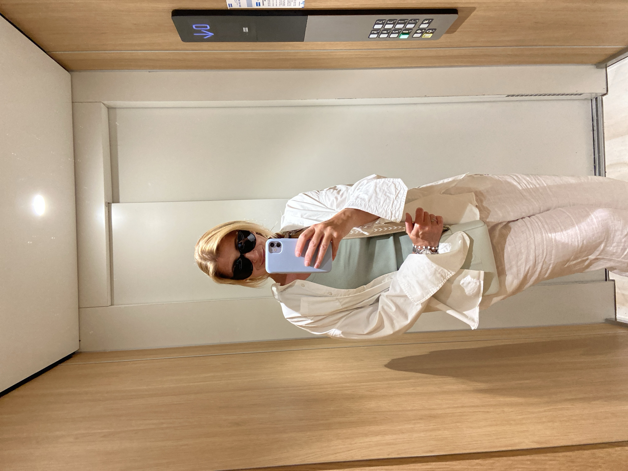 A woman captures a mirror selfie inside a modern elevator, likely located in Budva, Montenegro. The blonde-haired woman, wearing dark sunglasses, is dressed in a stylish light-colored ensemble featuring a long-sleeved button-up shirt, wide-leg trousers, and a light green top underneath. She holds a light blue smartphone in her right hand to take the picture, and a light green handbag is visible near her left arm. A silver-toned watch adorns her left wrist.
The elevator interior is characterized by light wooden paneling on the floor and ceiling, with smooth, light-colored walls. An elevator control panel is mounted on the ceiling, clearly displaying "01" along with an upward arrow, indicating the current floor and direction of travel. Below this, a grid of numbered buttons is visible, with several illuminated in green, suggesting selected floors. A small, illegible label with text is also present above the main control panel. The wooden floor reflects the woman's shadow and a partial view of her legs, adding depth to the image. The scene suggests a clean, well-maintained, and contemporary environment.