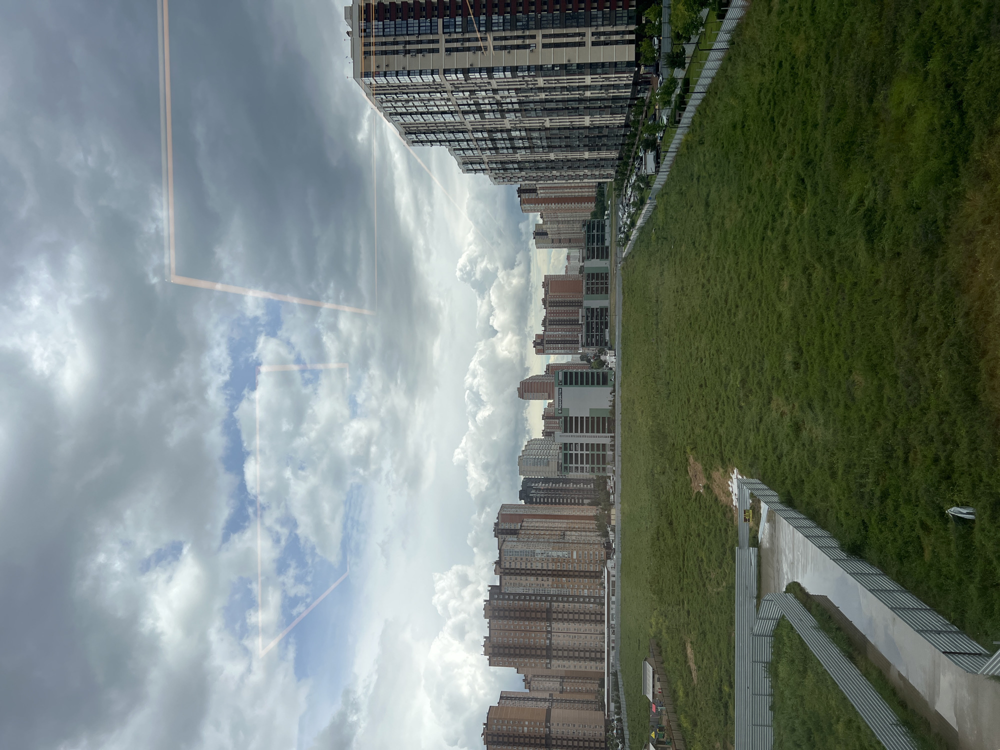 The image depicts a view of a cityscape, likely taken from a high vantage point within a building, given the presence of a window reflection. The location appears to be Krasnodar, Russia, based on the distinctive architectural style and urban planning.
The scene is divided between the sky, the buildings, and a green area, possibly a park or large lawn. The sky is filled with cumulus clouds, ranging from dense and dark to lighter and more dispersed.
The buildings form the central band of the image. They are modern, high-rise residential structures, with varied heights and façade designs, primarily in shades of brown and grey.
The green area occupies the lower portion of the view. The grass is long and dense. There is a metal walkway or path with a railing running alongside it.