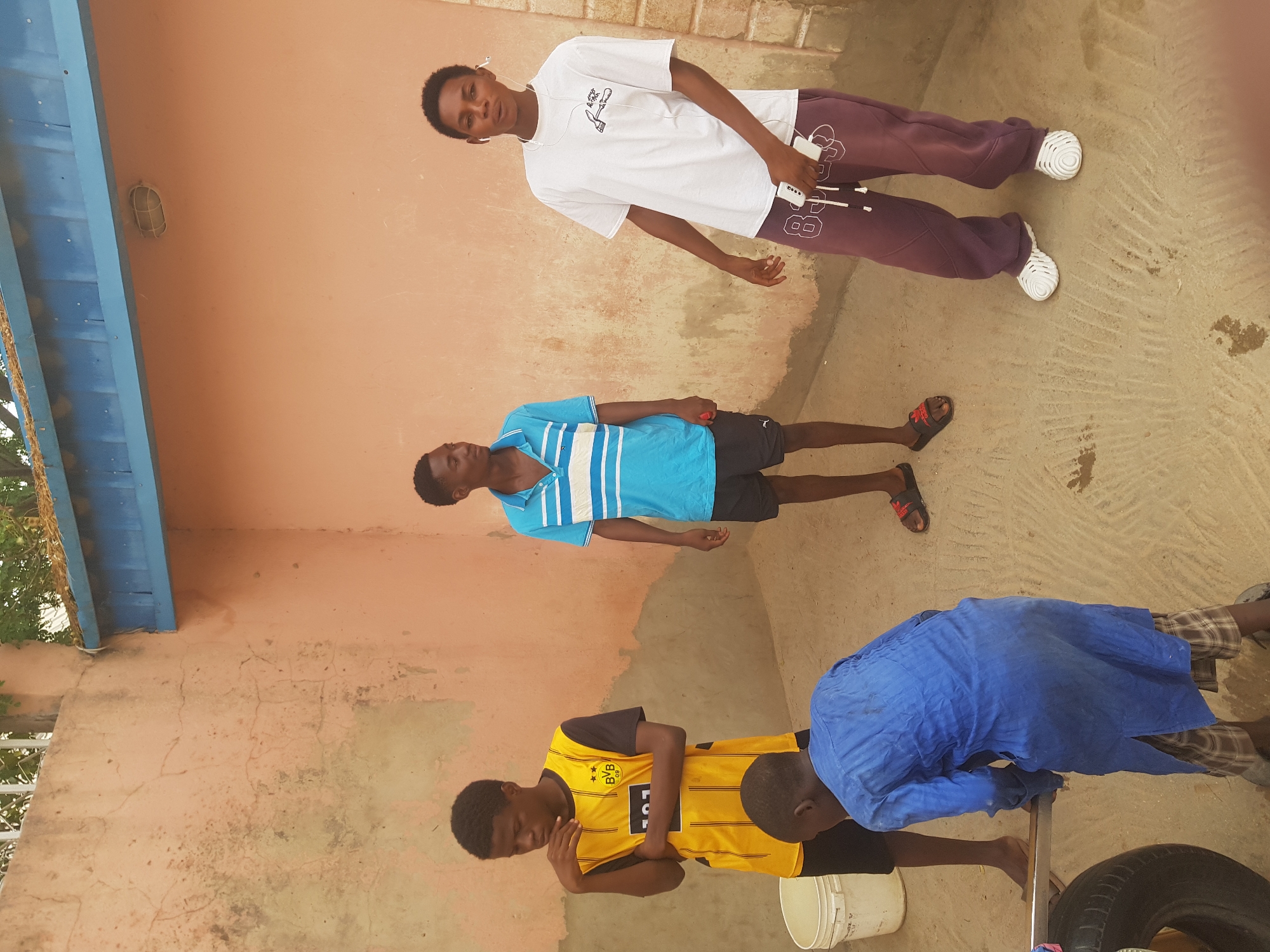 The image shows a group of four young men standing in an outdoor space in Dutsinma A, Nigeria. One is wearing a white t-shirt, maroon sweatpants and white shoes. The second individual is wearing a light blue polo shirt with white stripes and black shorts. The third is in a yellow soccer jersey. The fourth man, wearing a dark blue coverall, appears to be working on a car tire with a metal tool. A white bucket is near the tire. They are positioned on a concrete surface adjacent to a peach-colored wall. A blue awning is partially visible in the background.