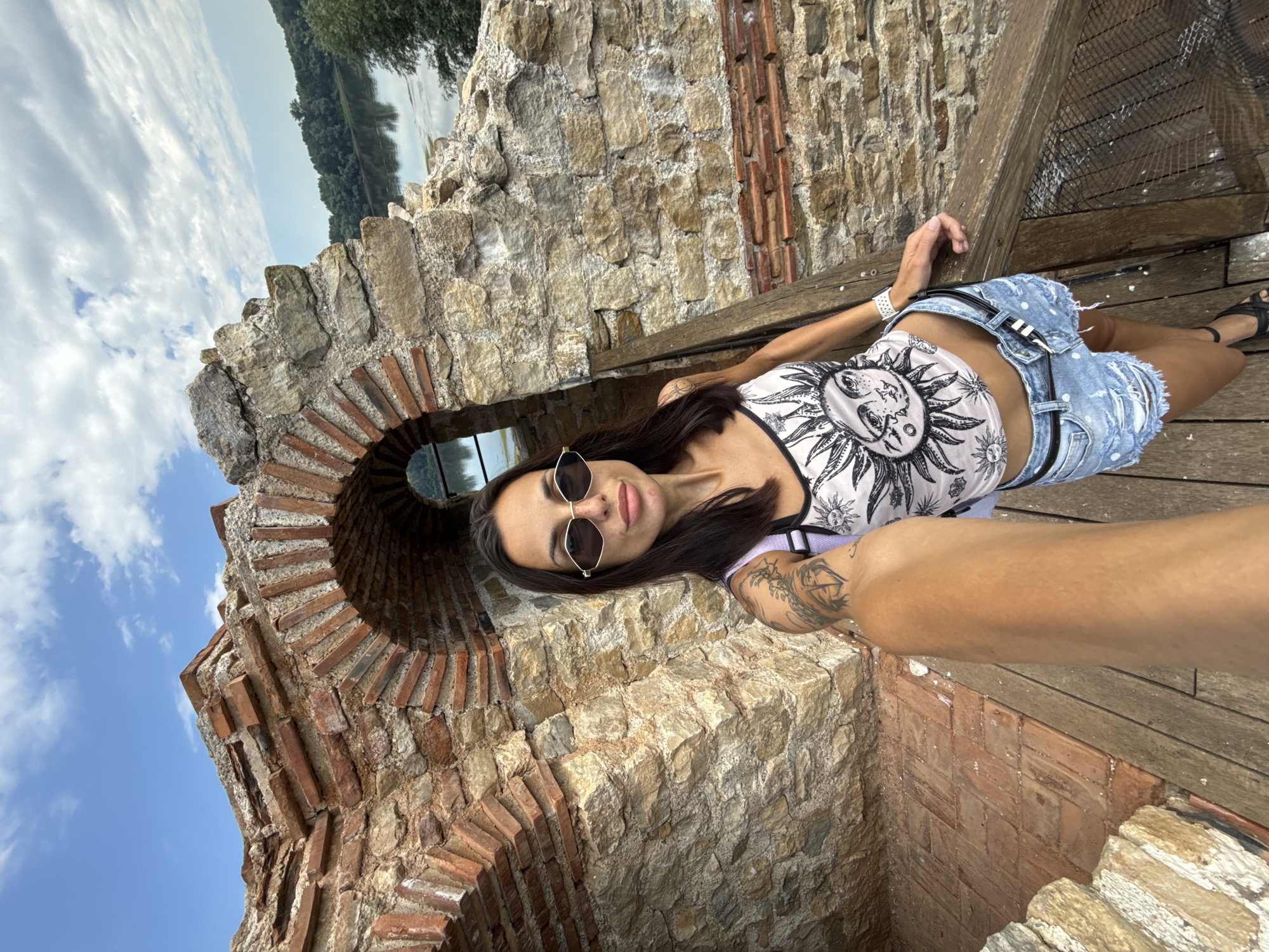 A woman with dark hair and sunglasses takes a selfie in front of ancient stone ruins. She is wearing a white tank top with a sun graphic, ripped denim shorts, and black sandals. She has tattoos on her left arm. The ruins are made of large stones and red bricks, with an arched opening. Beyond the ruins, a body of water, trees, and a cloudy sky are visible. The scene appears to be outdoors, likely at a historical site. The location is identified as the Fortress of Belene in Belene, Bulgaria.