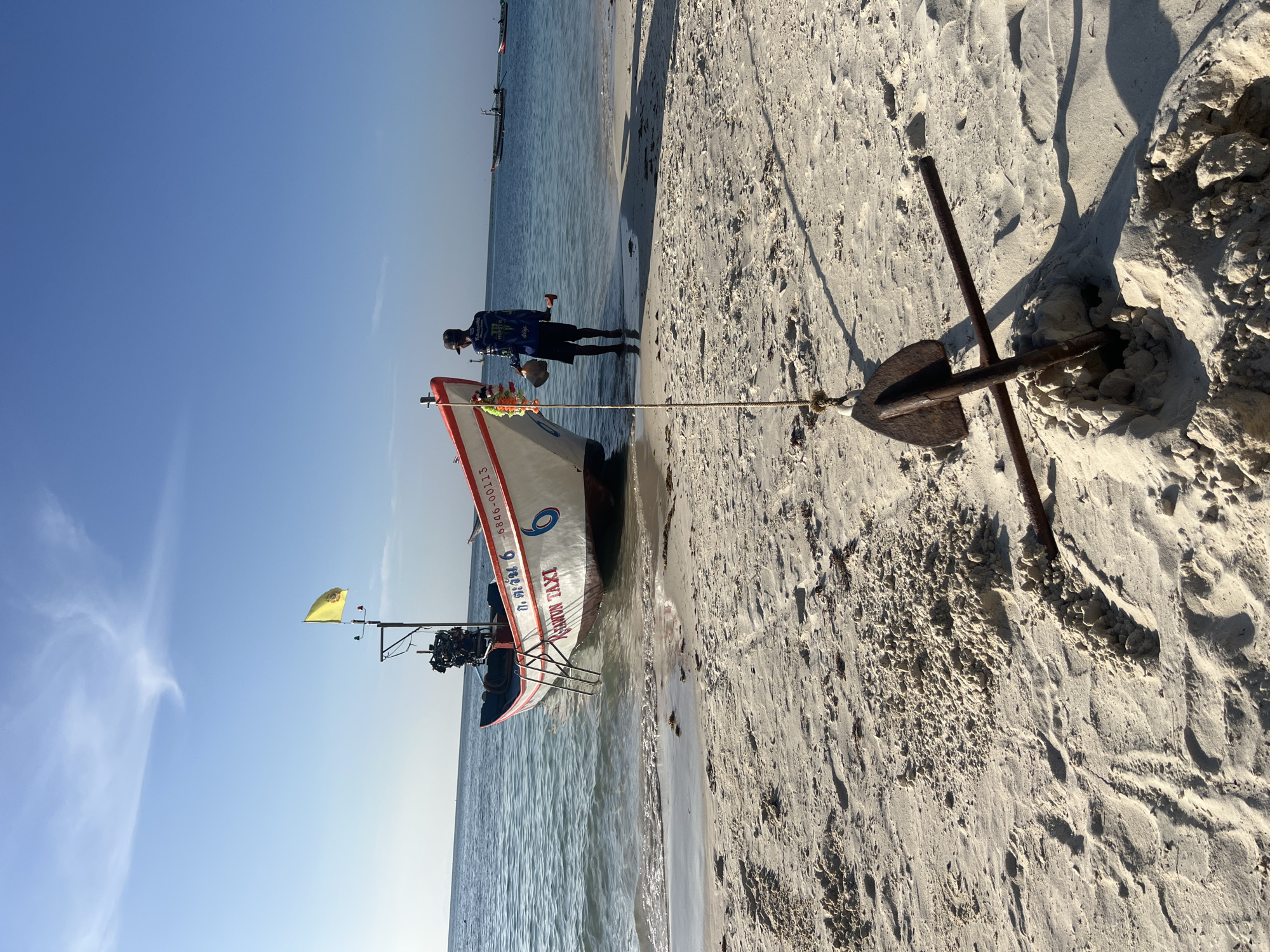 A medium shot shows a sandy beach with calm blue water lapping at the shore in Ko Pha-ngan District, Thailand. In the foreground, an anchor is partially buried in the sand, connected to a rope. A small boat with a yellow flag, an outboard motor, and the words "CHANUN TAXI" visible on its side, is beached in the shallow water. A person wearing a blue shirt and black shorts stands near the boat, holding a coconut and a tool. Another boat is visible in the distance on the water. The sky is clear and blue with a few wispy clouds.