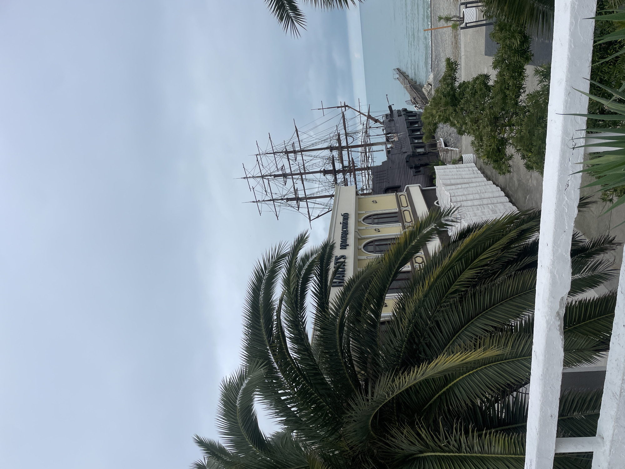 The image captures a view of the "Harat's Krasnodar" restaurant in Sochi, Russia. The restaurant building has a pale yellow facade, with its name displayed in dark blue lettering. Above the building, there's a large, non-functional pirate ship structure, primarily black with prominent masts and rigging. The sky is overcast and gray. In the distance, the sea is visible, with a breakwater or pier extending into the water. A palm tree is featured prominently in the foreground, partially obscuring the view. A white painted railing is partially visible in the lower right.