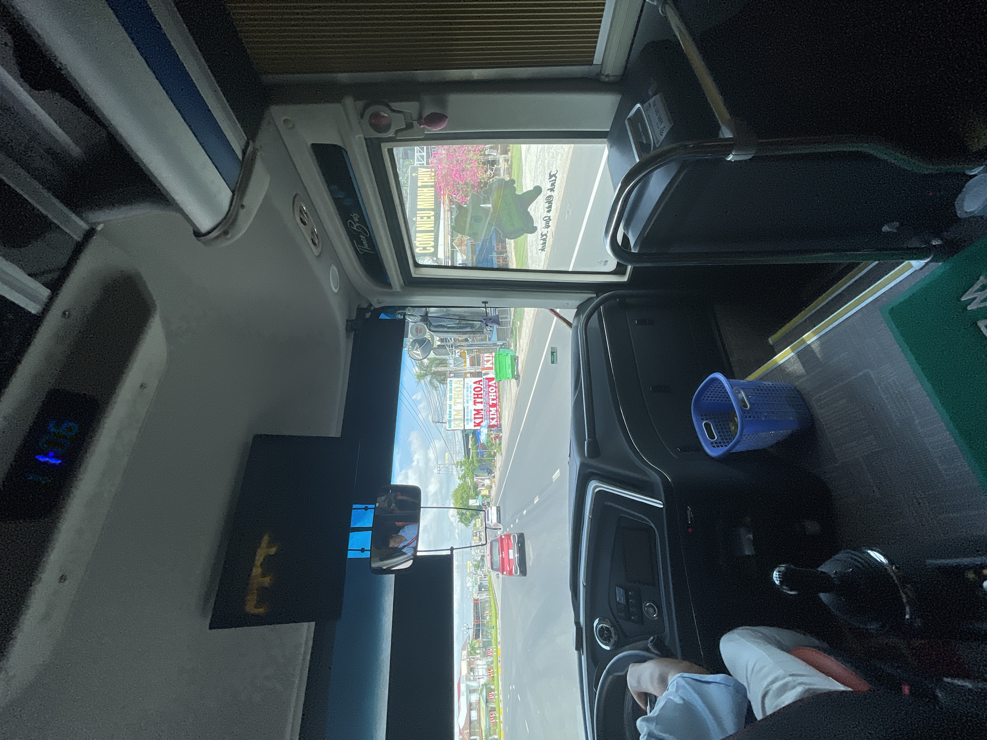 The image is taken from the interior of a bus, looking forward through the windshield. The bus appears to be in motion on a road, with other vehicles visible. The dashboard and steering wheel are prominent in the foreground, with a driver's hand on the wheel. To the right, a window shows scenery outside, including buildings, trees, and a pink flowering tree. Signs with Vietnamese text are visible on the roadside. Inside the bus, a blue plastic basket is on the floor, and a metal handrail is present. A digital clock on the ceiling displays "11:06". The overall impression is a snapshot of a bus journey in a Vietnamese city.