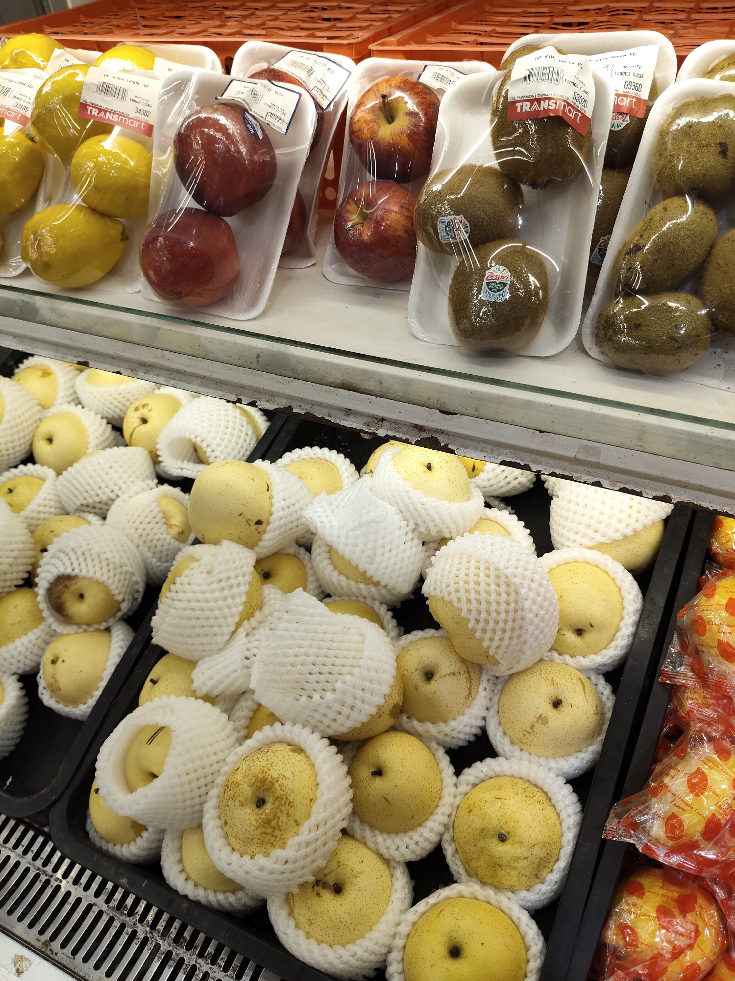 The image displays a retail display of fruits in a supermarket in Medan, Indonesia. In the foreground, several crates are filled with yellow pears, each individually wrapped in white foam netting. The pears are round and have a light yellow skin with some brown speckling. Behind the pears, on a higher shelf, are packages of red apples and kiwis. The apples are a deep red color and are also individually packaged. The kiwis are brown and fuzzy, packaged in white trays. Further in the background, a shelf holds more yellow lemons, also packaged in white trays. The overall scene is a well-stocked fruit section of a grocery store, with a focus on fresh produce.