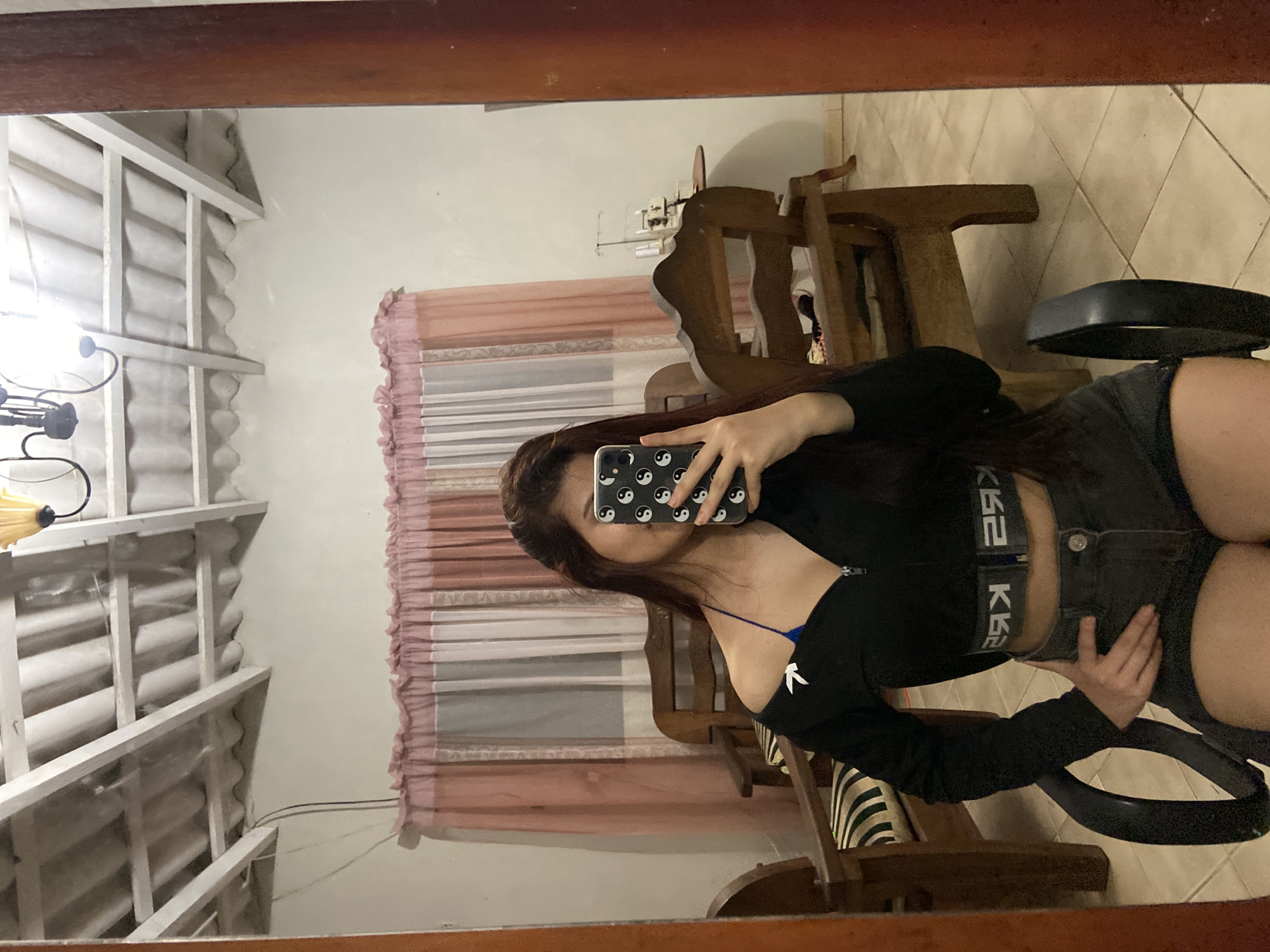 The image is a mirror selfie of a woman. She is holding a phone with a yin-yang patterned case. The woman is wearing a black, long-sleeved crop top with a zipper and dark shorts with a black waistband. The room is interior with pink curtains, wooden furniture, and a decorative chandelier. The location is not specified in the image and cannot be determined.