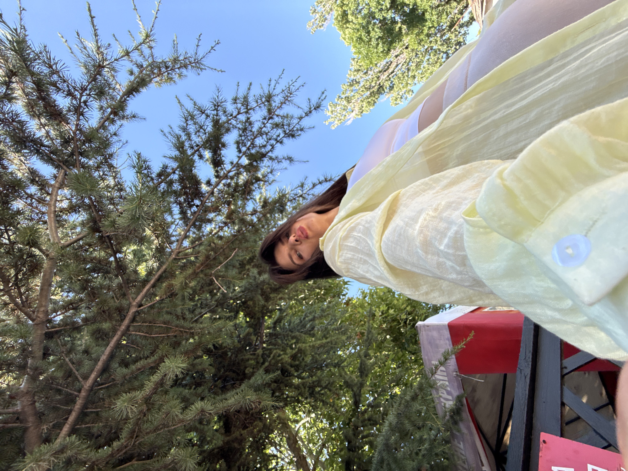 This is a low-angle, wide shot of a person outdoors. The person, identified as a young woman, is wearing white underwear and a light yellow, translucent shirt. Her face is visible, looking towards the camera, with a relaxed expression. The scene is set amongst trees with lush green foliage and a clear blue sky visible. Sunlight suggests it is daytime. A part of a structure, possibly a tent or gazebo with red and white fabric, is visible in the background, along with some wooden supports. The overall impression is of a casual, outdoor setting.