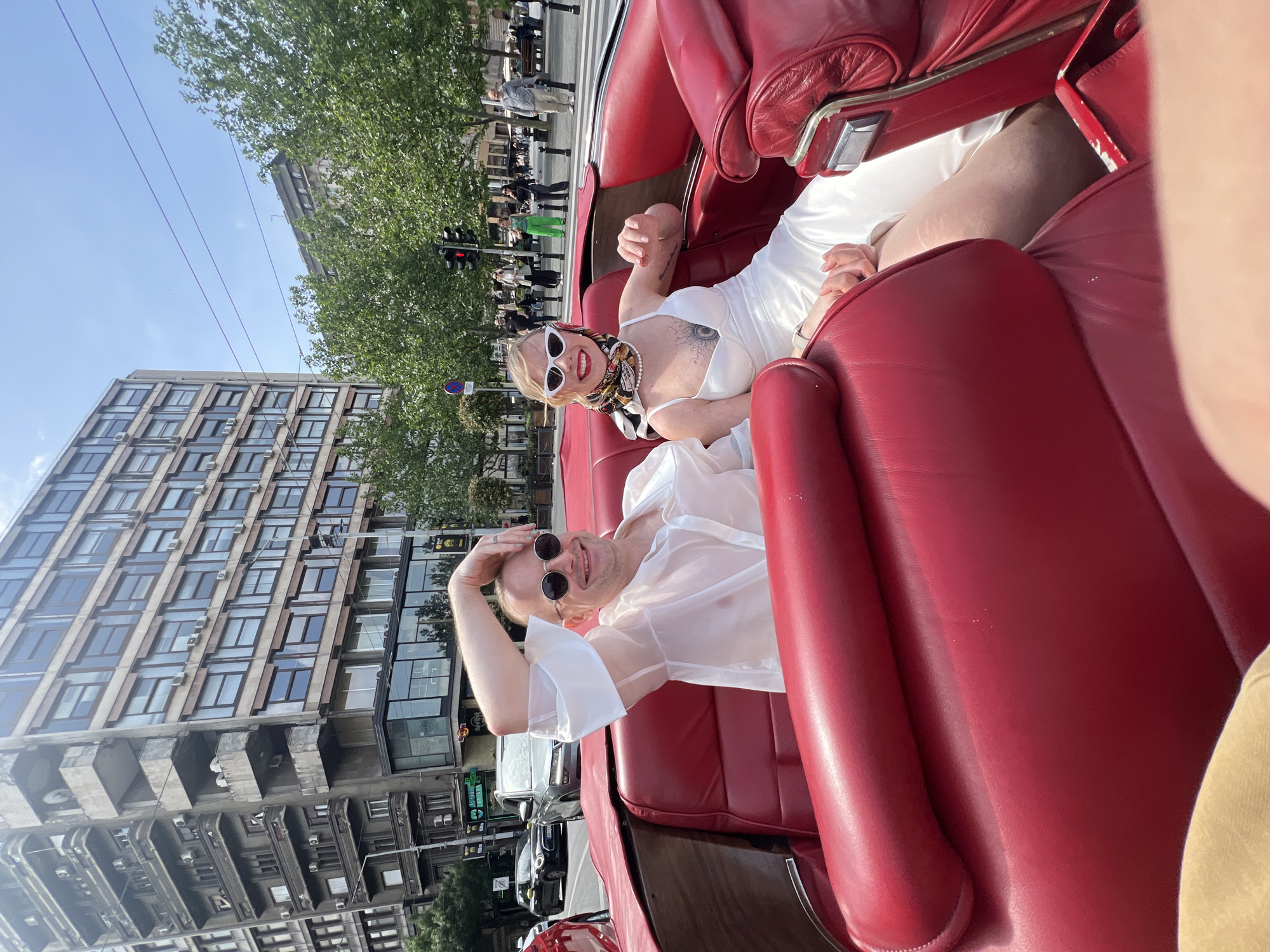 A medium-shot, slightly rotated, outdoor photograph shows two people in a red, convertible car. In the front passenger seat, a woman with blonde hair and white sunglasses, wearing a white dress, a scarf around her neck, and a pearl necklace, is smiling and gesturing with her left hand. In the back seat, a man with short hair and sunglasses, wearing a sheer white shirt, is smiling and holding his hand on his forehead. The car is stopped at a crosswalk in a city. Buildings and trees are in the background. Traffic lights are visible, and pedestrians are on the sidewalk. The photograph was taken in Belgrade, Serbia.