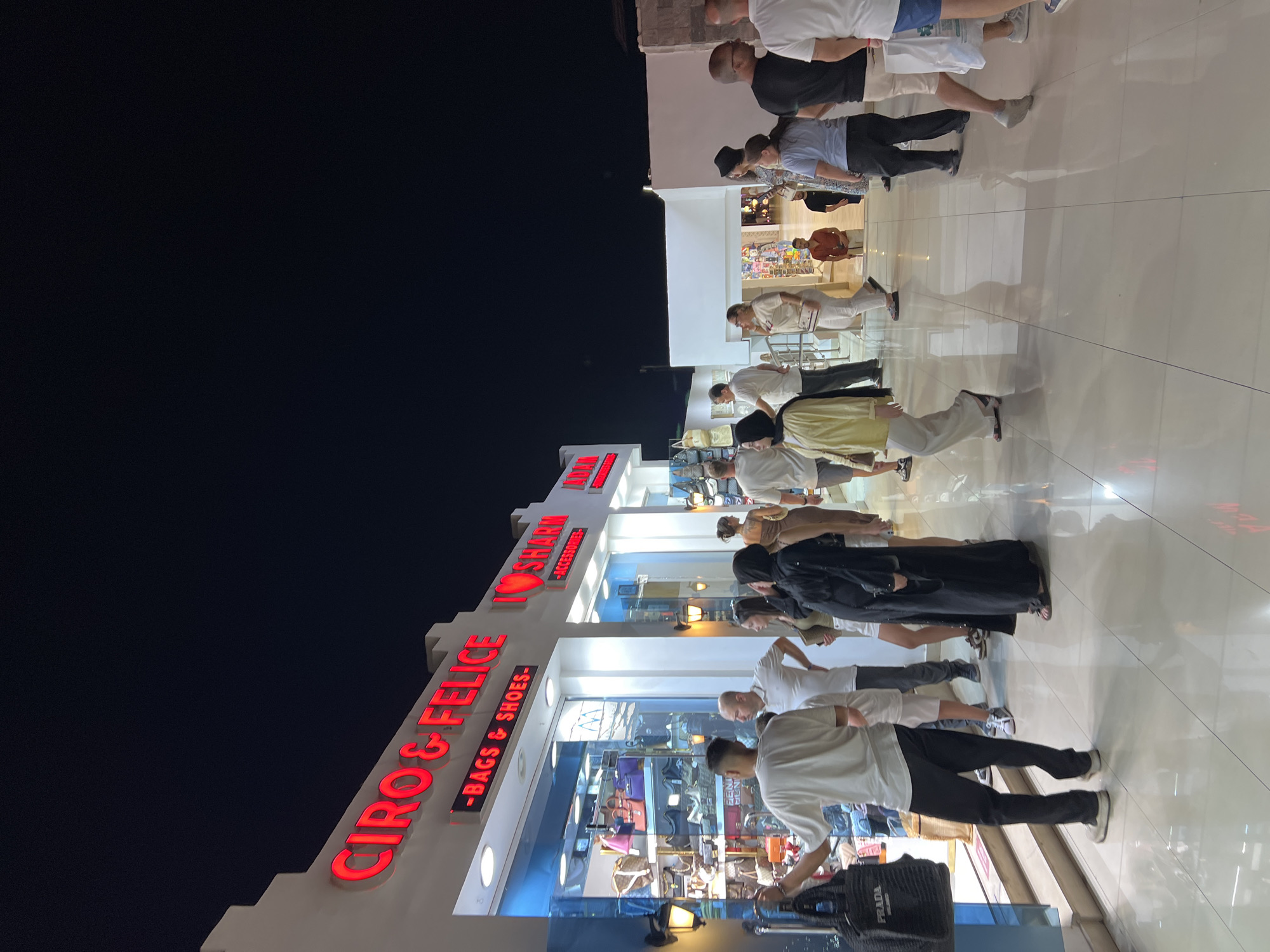 The image depicts a nighttime scene in Sharm El Sheikh, Egypt, likely on a promenade or shopping street. The foreground is dominated by a group of people walking and standing on a polished tiled floor. Several individuals are dressed in casual attire.
To the left, a store with the prominent red neon sign "CIRO & FELICE - BAGS & SHOES -" is visible, with its interior displaying various handbags and shoes. Another store to the right has a similar red neon sign that reads "I LOVE SHARM - ACCESSORIES -". The shops are illuminated, and external lights are also present.
In the middle ground, more people are seen strolling. One person is wearing a yellow cardigan, and another is in a black abaya. Behind them, a man in a white polo shirt and another in a white t-shirt are interacting or observing.
The background shows more shops and open spaces, with the dark night sky overhead. The overall atmosphere suggests a lively commercial area in a tourist destination.
