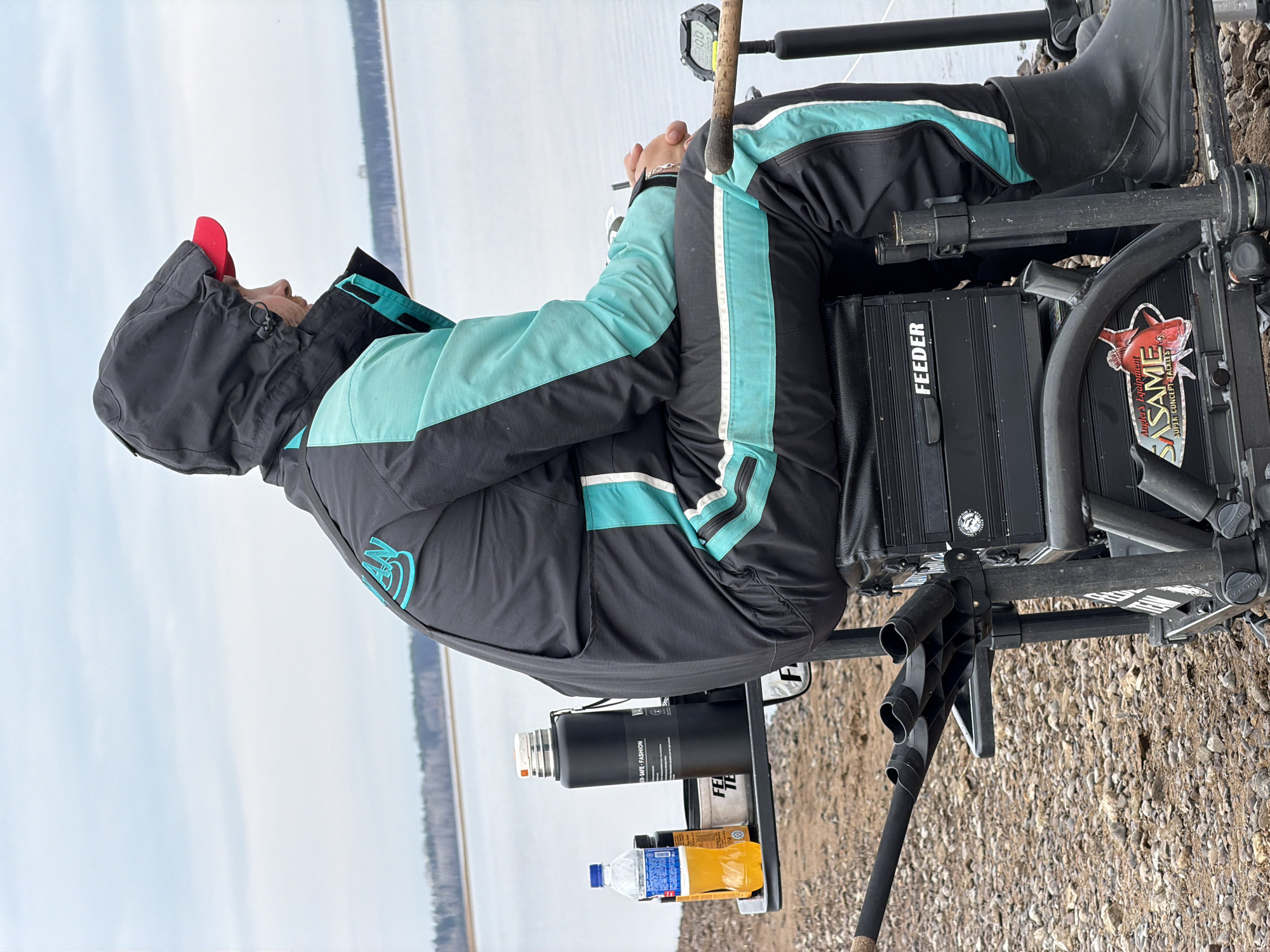 The image shows a person sitting in a fishing chair on the shore of a body of water, possibly the Kama Reservoir. The person is wearing a dark jacket with teal accents and a red cap, their face partially obscured by the hood. They are seated on a specialized fishing platform chair, which includes storage boxes labeled "FEEDER" and "SASAME," as well as attachments for holding fishing rods. There is a small table attached to the chair holding a thermos and a couple of beverage bottles. The background shows the calm water surface and the opposite shore with trees. The ground at the person's feet is gravelly. Given the name "Sasame" and based on the geographic location of the image, it's likely the city of Chaikovsky, Russia. The image is taken from a low angle.