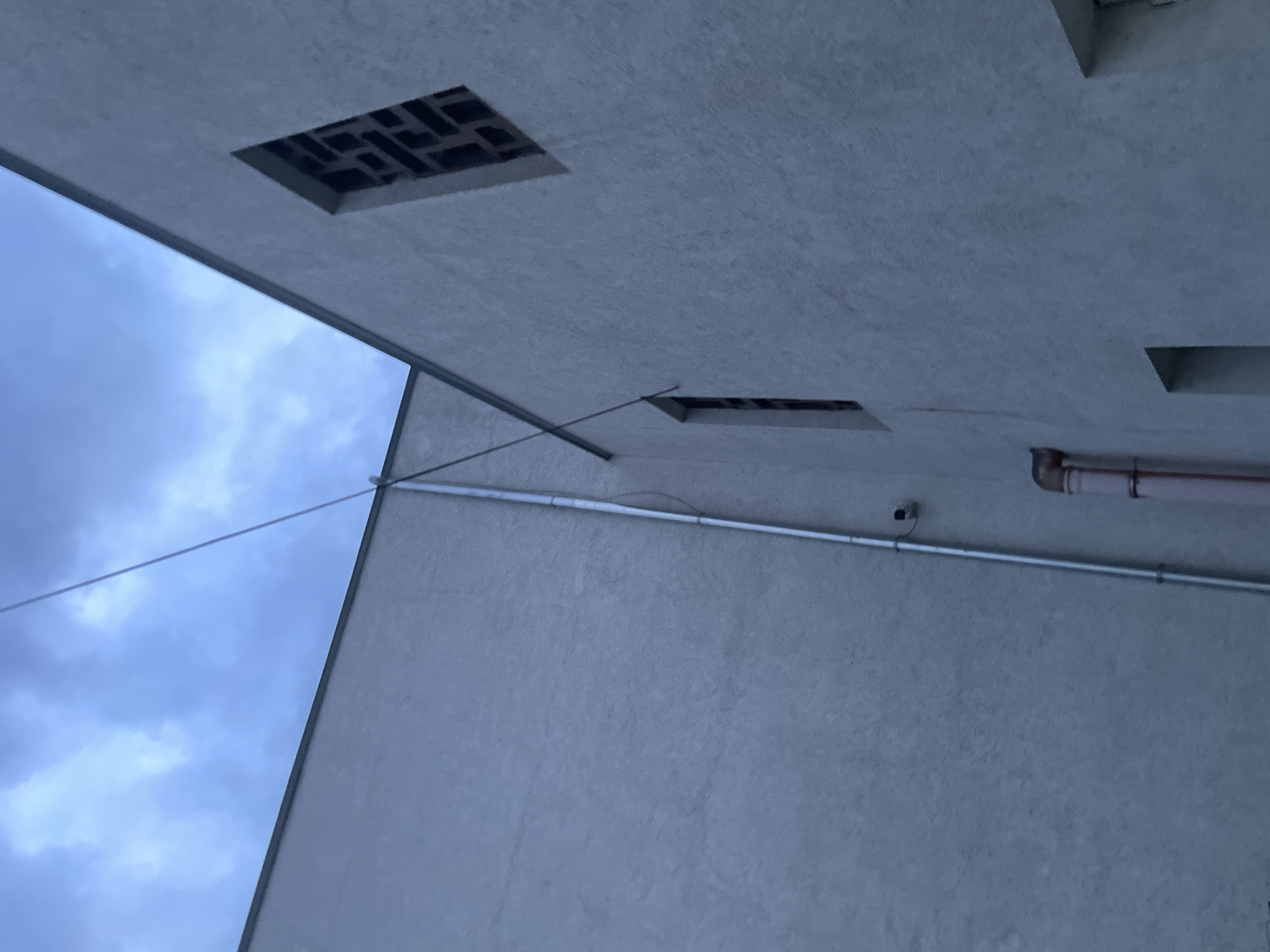 The image captures an exterior view of a building's architecture against an overcast sky, taken from a low and tilted perspective, looking upwards. The scene is dominated by two large, light-grey, textured surfaces of a building, possibly concrete or stucco, meeting at an angle in the upper-middle of the frame.
In the upper section, a wide, rectangular opening with a dark, geometric, grid-like pattern is visible within the building structure. Below and to the right of this, on a different building plane, a narrower, elongated rectangular opening also appears.
A thin, dark wire or cable diagonally crosses the image, stretching from near the top-center towards the lower-middle left, where it meets a thicker, horizontal conduit running across the lower building surface. Attached to this conduit, a small, dark, dome-shaped object, likely a security camera or sensor, points outwards. Further to the right, a system of larger, reddish-brown pipes, possibly for drainage or other utilities, is visible, featuring an elbow joint and mounting straps.
The upper-left portion of the image reveals a pale blue sky filled with soft, diffused white and grey clouds, indicating an overcast daytime. The overall lighting is dim and soft, with no harsh shadows. No people or text are visible. The setting is an outdoor area in Ihtiman, Bulgaria.