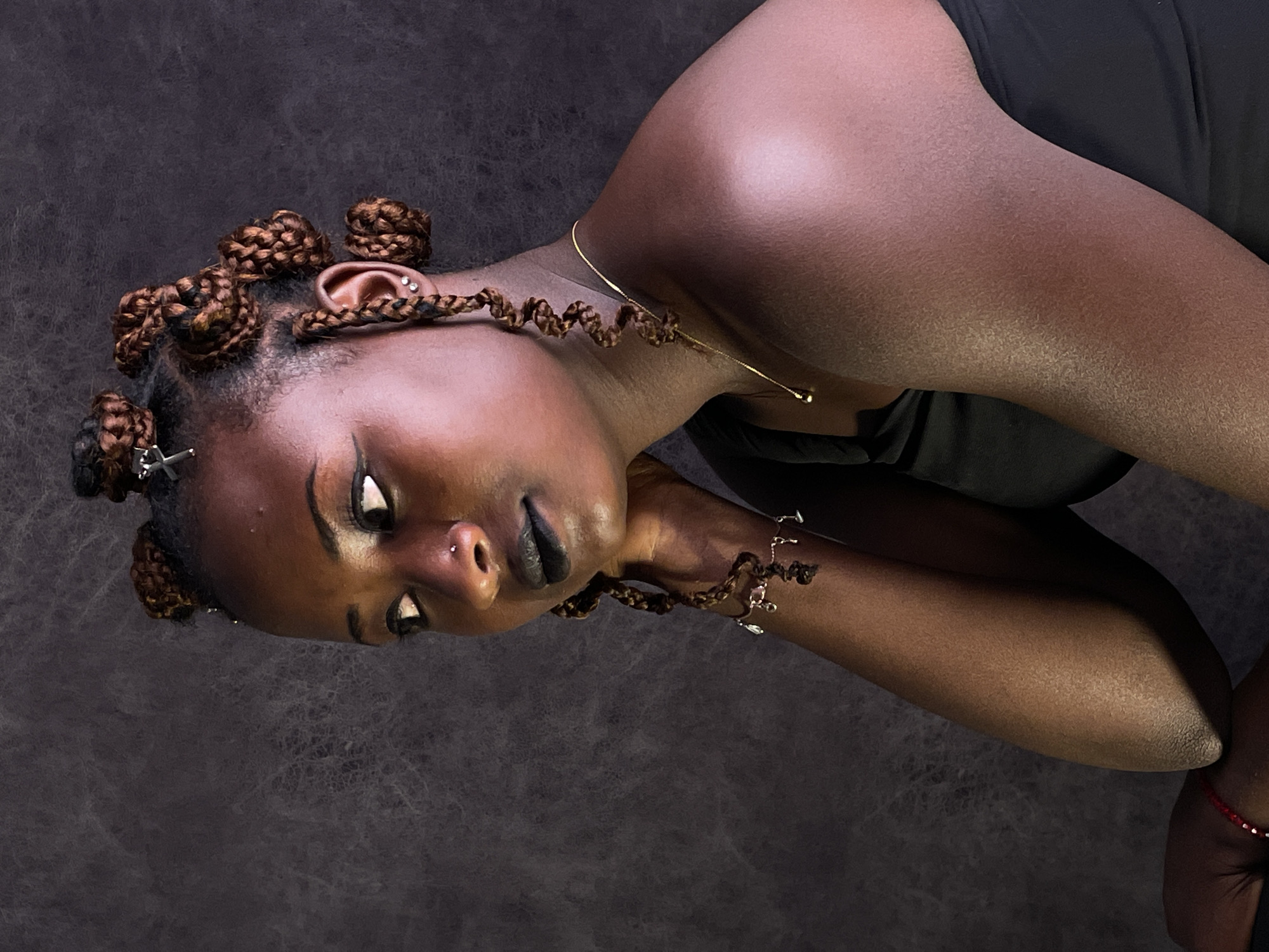 A close-up, rotated portrait features a dark-skinned person with their head tilted, suggesting a reclining or relaxed pose. The individual's face is prominent, with eyes partially closed and looking downwards, conveying a serene or contemplative expression. Dark lipstick adorns their full lips, and a small stud is visible in a nose piercing.
Their hair is styled in intricate, reddish-brown braids fashioned into several tight, round buns (likely Bantu knots) across the scalp. Some braids extend loosely, decorated with small metallic accessories. The person wears multiple pieces of jewelry, including two stud earrings on their visible ear, a larger decorative earring on the side of their head, a thin gold chain necklace, and a silver-toned charm bracelet on their left wrist. A red string bracelet is also visible on their right wrist.
The individual is wearing a dark, possibly black, garment that covers the torso. The background is a uniformly dark, textured surface, typical of a studio backdrop, which serves to highlight the subject. The lighting is soft, creating gentle highlights on the skin, particularly on the shoulder and cheekbone. The image captures a moment of quiet introspection or calm during a photographic session. There is no visible text in the image. The provided context indicates the location is Barakin Akawo, Nigeria.