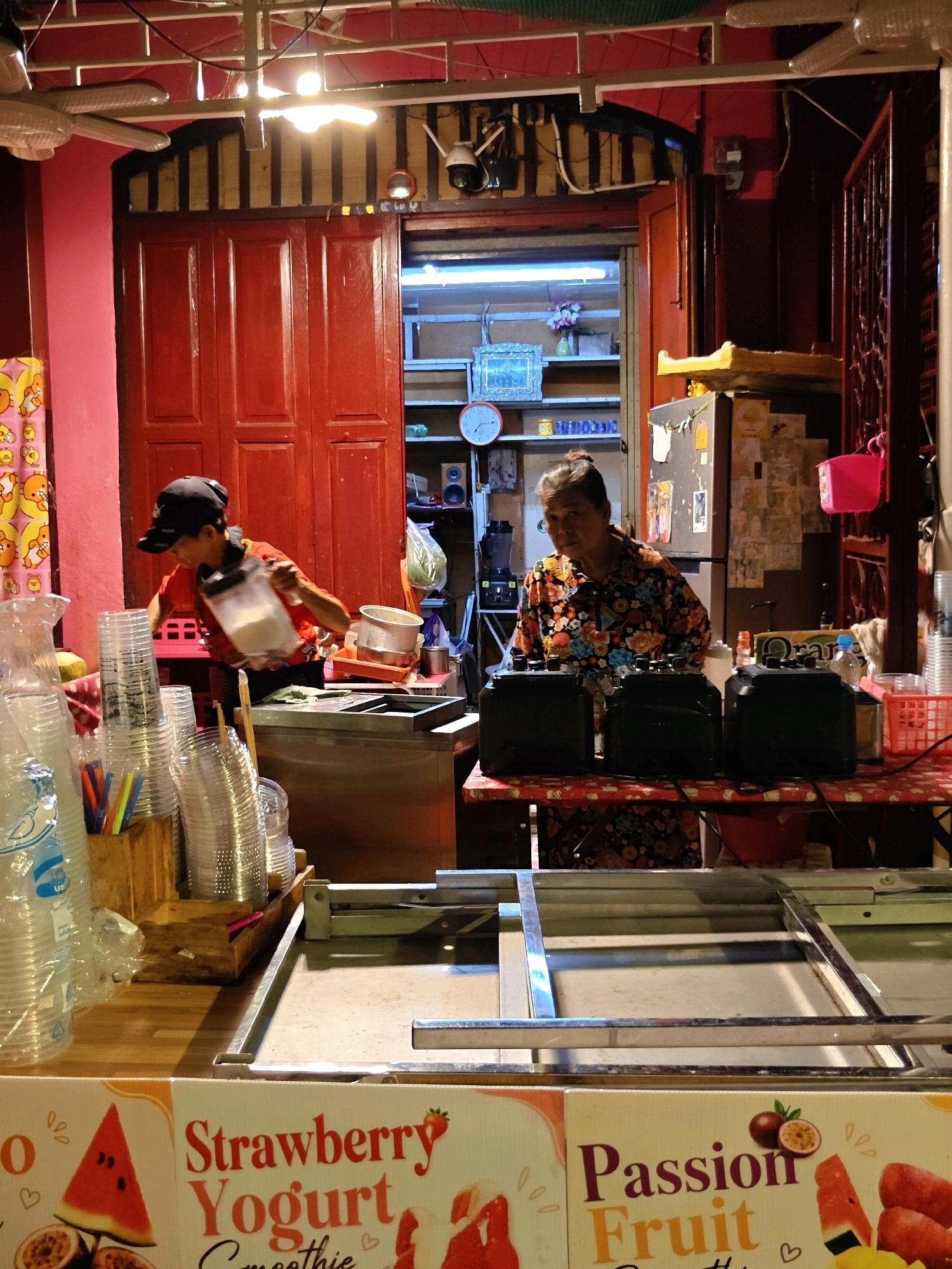 The image depicts a street food stall in Bangkok, Thailand. In the foreground, there is a metal preparation counter with various plastic cups and straws neatly arranged on the left. Behind the counter, a woman in a floral-patterned shirt is standing, looking towards the camera. To her left, another person wearing a black cap and an orange shirt is actively preparing something, holding a clear plastic container. The stall features a rustic wooden door in the background, with shelves and appliances visible beyond it. The overall setting suggests a busy, informal food vending operation typical of Bangkok's vibrant street culture.