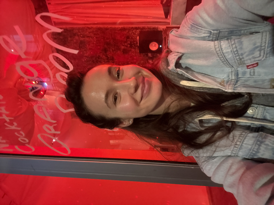 A woman smiles in front of a red-lit window. "Cocktails or Wine" is written in white on the window, along with a disco ball. The woman wears a light blue denim jacket over a gray hooded sweatshirt. The scene is likely indoors, possibly a bar or restaurant with vibrant, modern lighting. A black device is mounted on the wall above her head.