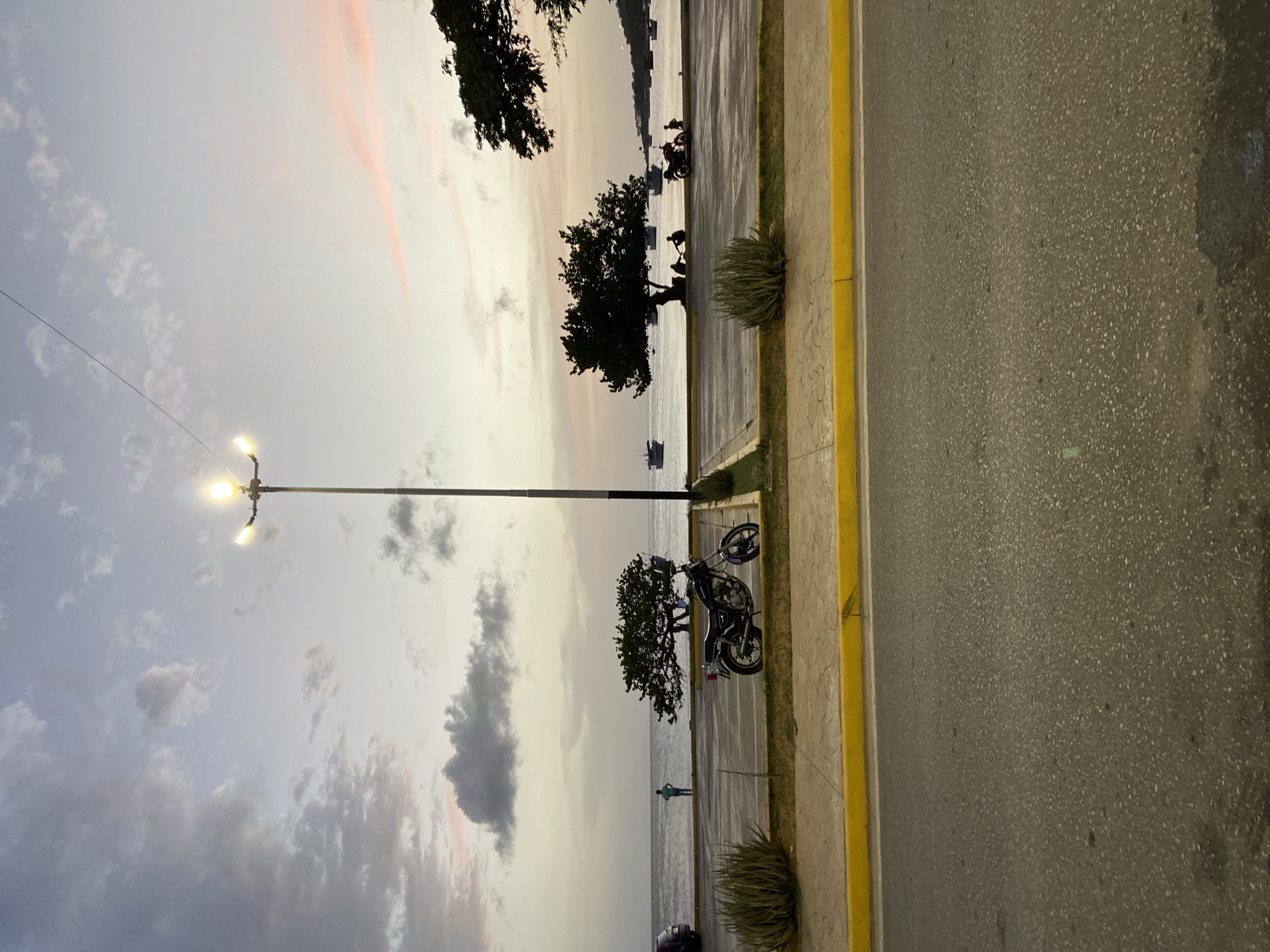 The image shows a twilight scene at the waterfront in Juan Griego, Venezuela. The sky is a gradient of pale blue and pink hues with scattered clouds. A tall lamppost with two illuminated bulbs stands prominently in the mid-ground. In the foreground, a paved road is visible with a yellow curb and a textured sidewalk. Beyond the sidewalk, a parking area contains a parked motorcycle and some arid, spiky plants. The middle ground features the Caribbean Sea with several boats docked or anchored offshore. On the shoreline, a lone figure stands looking out at the water, and further along, silhouettes of people are gathered near a tree. The overall atmosphere is serene and tranquil, characteristic of a coastal town at dusk.