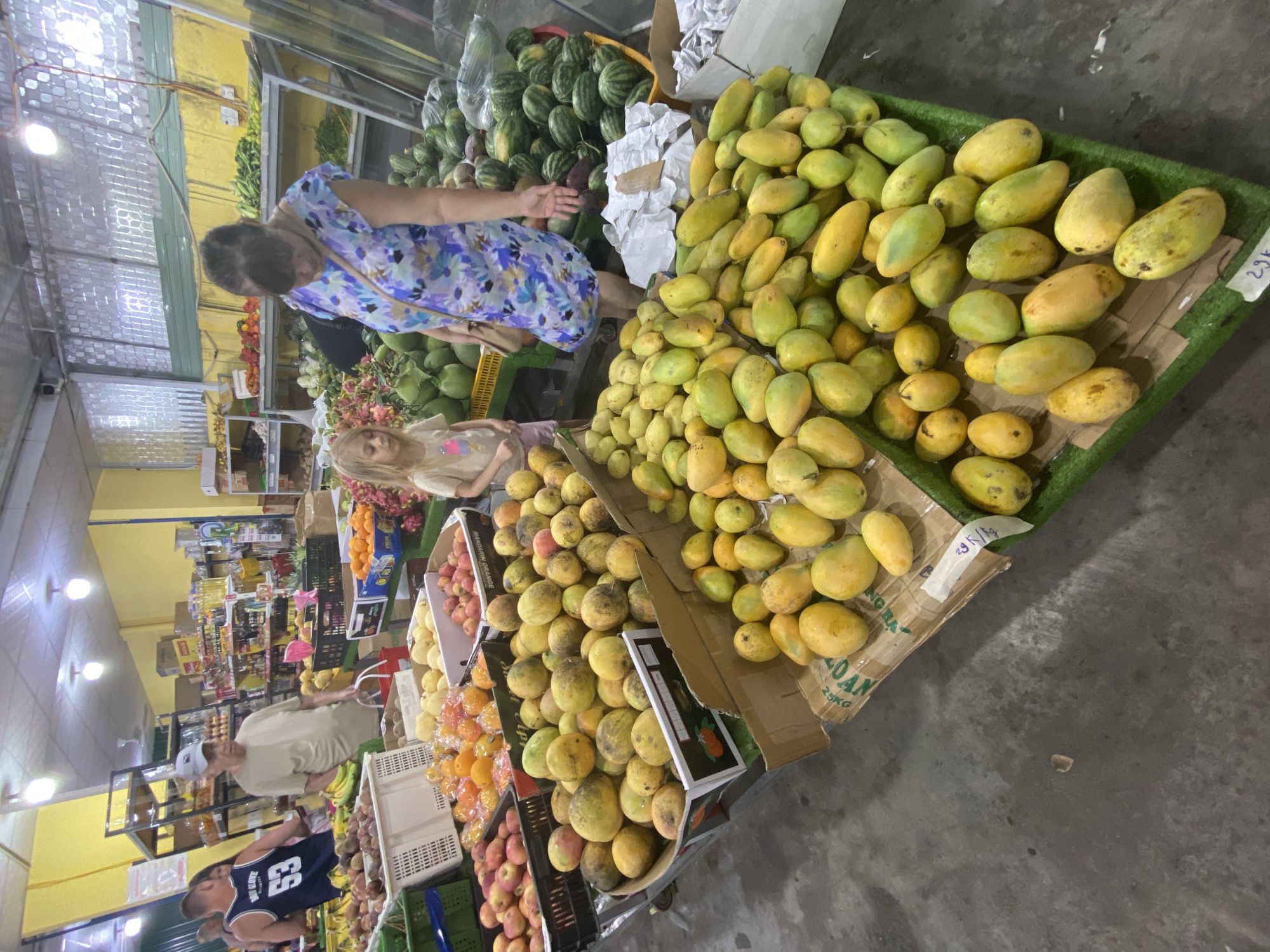 A fruit market stall in Bac Nha Trang Ward, Vietnam, displays a variety of fresh produce. In the foreground, a large quantity of ripe yellow mangoes are piled high in cardboard boxes and on a green artificial turf display. Behind the mangoes, a woman in a blue floral dress is looking down at the fruit, while a young girl with blonde hair stands beside her, looking at the mangoes. To the left, a man in a navy blue tank top with the number "53" on it is partially visible. In the background, shelves are stocked with various fruits and vegetables, including watermelons, tomatoes, and what appear to be dragon fruits. The market appears to be indoors, with fluorescent lighting overhead and a concrete floor. The overall scene depicts a bustling market environment focused on the sale of fresh produce.