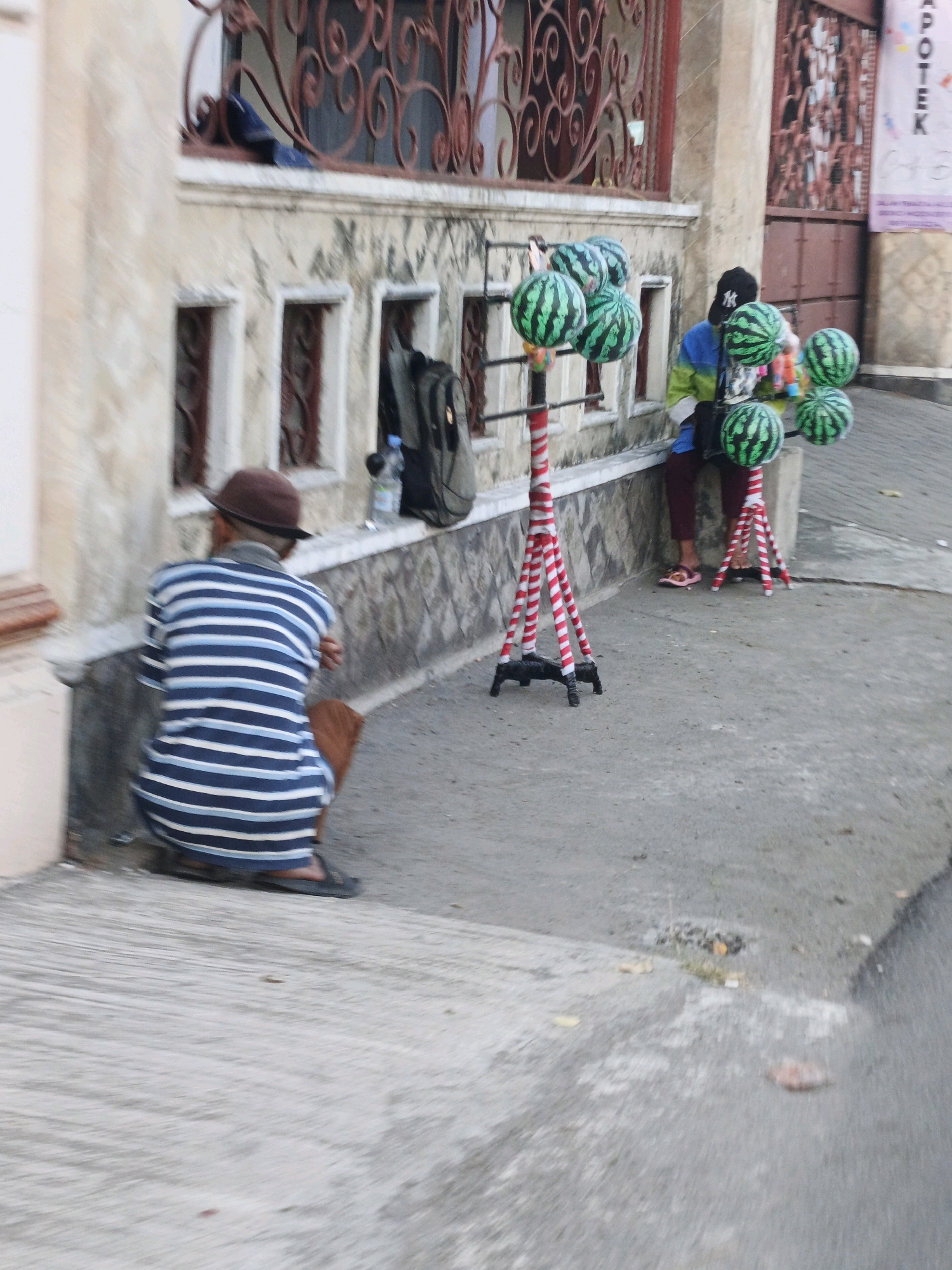 In the city of Sooko, Indonesia, a person wearing a blue and white striped shirt and a brown hat is crouching on the sidewalk. They are facing away from the camera, with their left arm resting on their leg. To their right, a person wearing a black baseball cap, a blue and green jacket, and dark pants is sitting on a concrete ledge. They are looking to their left. In front of the seated person, a display of several green and black striped inflatable watermelons is arranged on stands decorated with red and white stripes. One person is wearing pink sandals, while the other person's footwear is not clearly visible. Behind the seated individual, a backpack is leaning against a wall. The environment appears to be an outdoor setting with a building and a paved street.