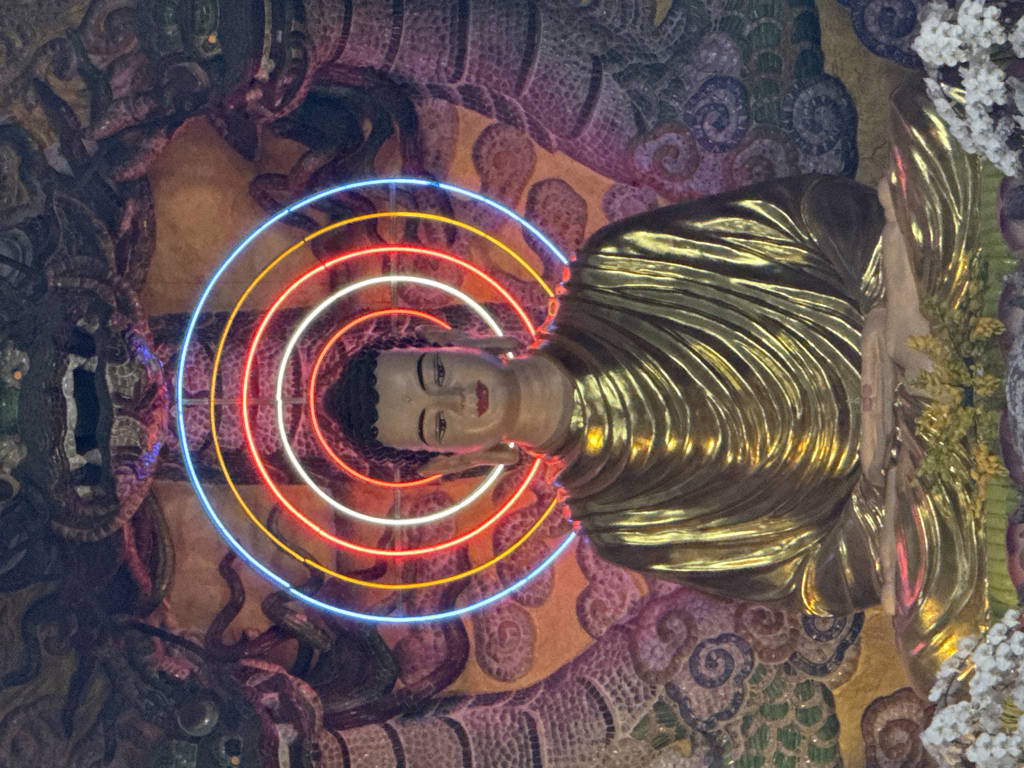 The image depicts a golden Buddha statue in a seated meditation pose, illuminated by a vibrant neon halo of blue, white, red, and yellow concentric circles behind its head. The backdrop is a richly textured, possibly mosaic or carved, wall featuring ornate dragon-like figures on either side, rendered in shades of pink, purple, orange, and green. The Buddha's robes are depicted with flowing, metallic folds, reflecting the ambient light. In the foreground, white and yellow floral arrangements are visible. The scene has an ethereal and spiritual ambiance, likely situated within a temple or shrine. This imagery is consistent with the artistic and architectural styles often found in religious sites within Lâm Viên - Đà Lạt, Vietnam, a region known for its Buddhist temples and pagodas.