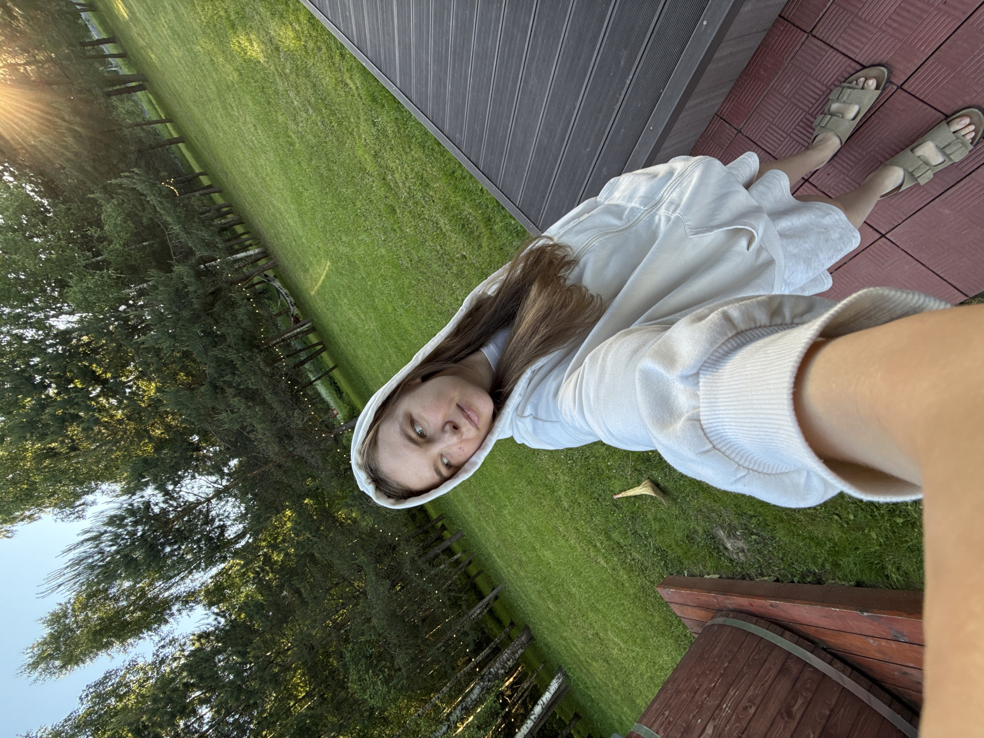 A young woman captures a low-angle selfie outdoors in Копнино, Russia, under bright, warm sunlight with a visible lens flare in the upper right. She is positioned on a reddish-brown tiled surface, adjacent to a dark grey slatted composite or wooden deck that extends along the right side of the frame. Her left arm, holding the camera, is extended into the blurred foreground. She wears a light-colored hooded sweatshirt over a light-colored dress or long shirt and olive-green open-toed sandals. Her long, light brown hair is visible beneath the hood. Her gaze is directed off to her left (viewer's left), appearing pensive. The background consists of a well-maintained, vibrant green lawn sloping gently upwards, bordered by a dense line of trees including birches with white bark and various evergreens. Decorative string lights are strung between some of the trees in the middle ground. A light-colored, natural-fiber broom-like object lies on the grass near a reddish-brown wooden platform in the left foreground. A large, dark brown wooden barrel with two metal bands rests vertically on this platform.